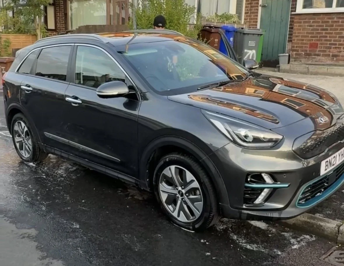 A dark gray Ford Kuga parked on a wet street in front of a brick house with garbage bins nearby.