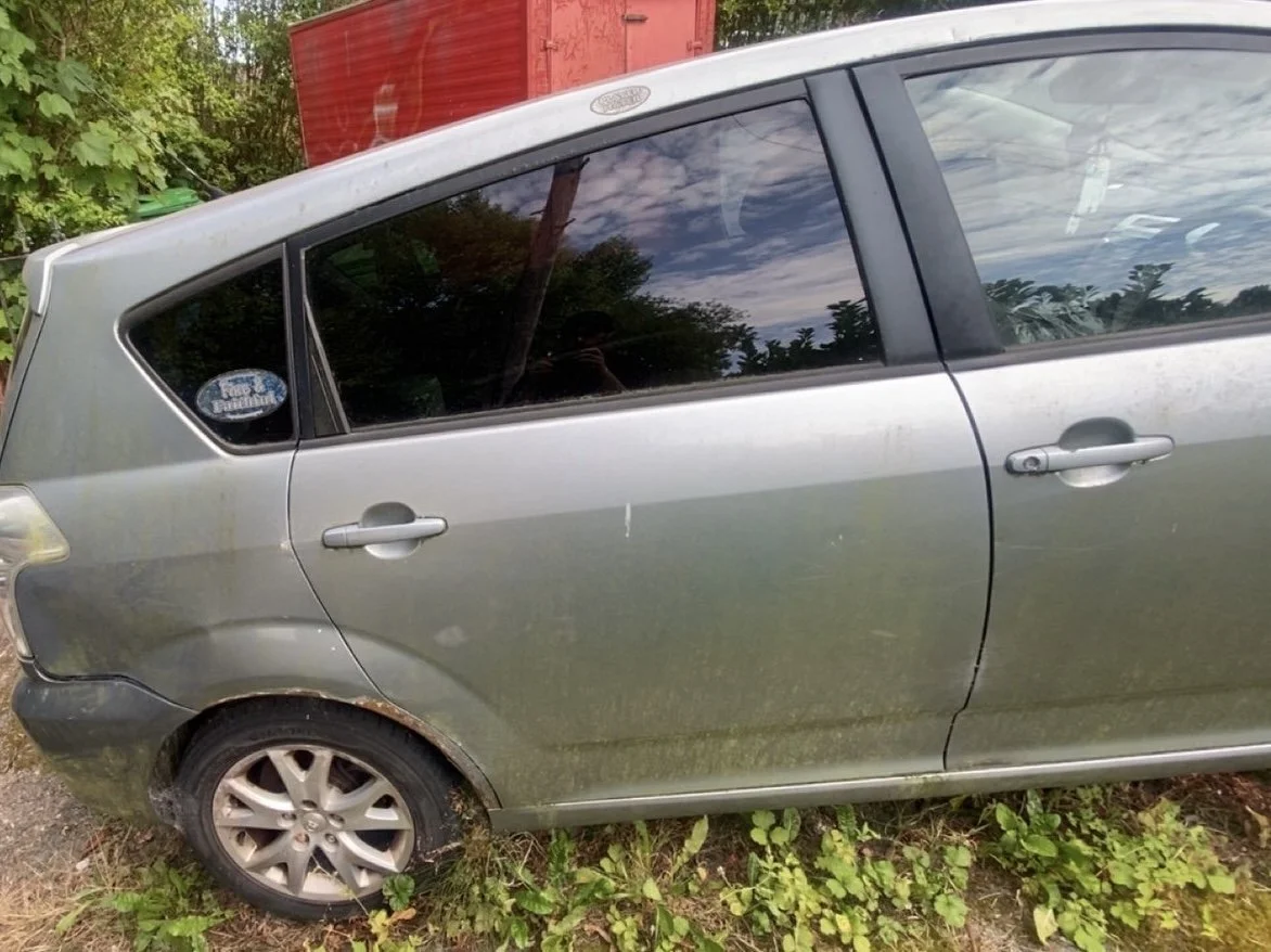 A silver hatchback car parked outdoors with overgrown grass and plants around the tires, which are dirty and appear slightly flat. The car is weathered, with visible dirt and minor scratches, and a parking sticker is on the rear passenger window.