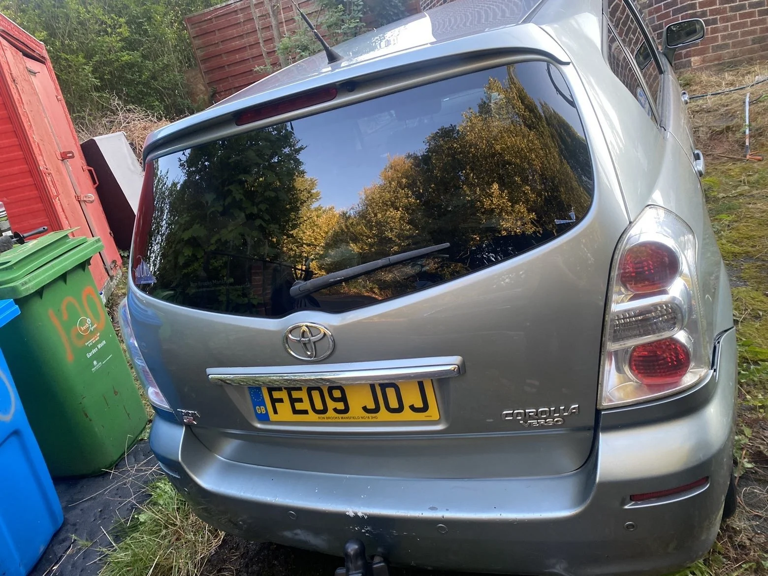 A silver Toyota Corolla Verso with a UK license plate FE09 JJJ parked outdoors near a brick wall, with trees reflected in the rear window and green and blue trash bins on the left.