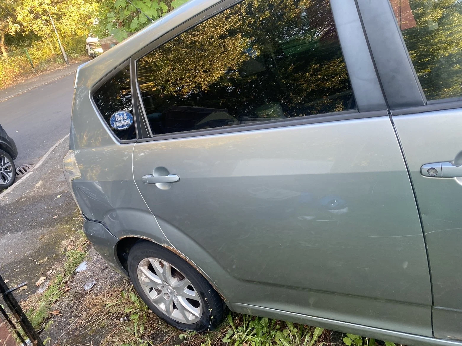 A silver hatchback car with visible damage on the rear passenger side, parked on the side of a street with trees and other vehicles in the background.