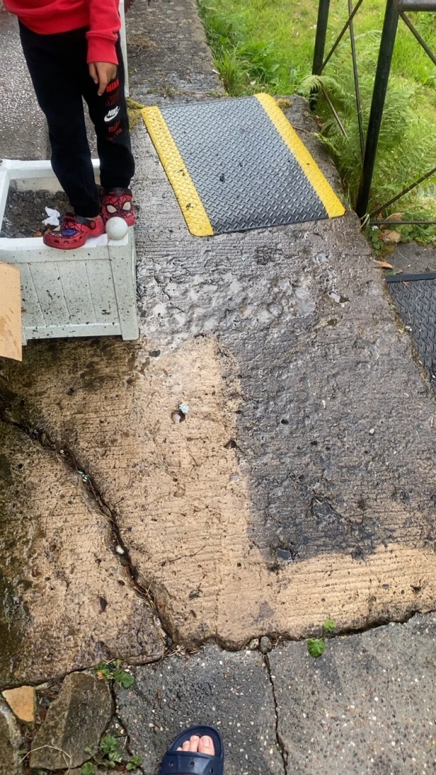 A person standing on a concrete bridge with visible cracks and wear, wearing black sweatpants and red sandals, next to a white planter and a golf ball, with a metal ramp on the sidewalk and lush green foliage nearby.