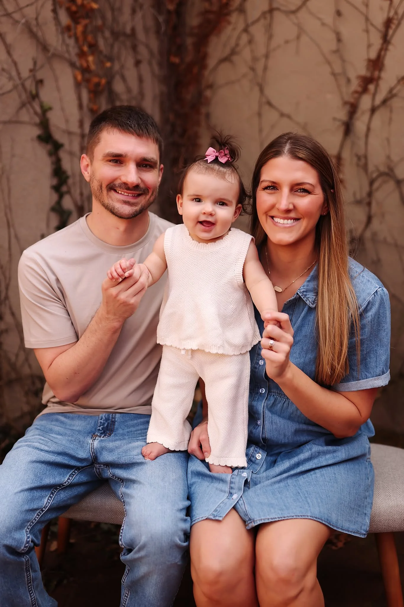 A happy family of three posing outdoors in front of a tree with vines. The man is on the left wearing a beige t-shirt and jeans, the woman is on the right wearing a denim shirt and shorts, and their young daughter with a pink bow in her hair is in the middle wearing a white sleeveless top and pants.