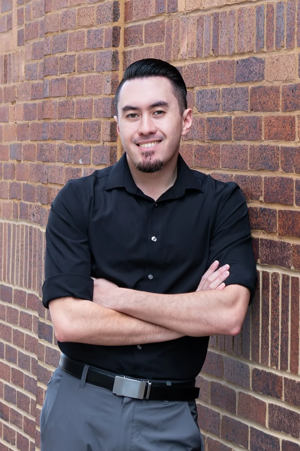 A young man with dark hair, a goatee, wearing a black button-up shirt and gray pants, standing against a brick wall with his arms crossed, smiling at the camera.