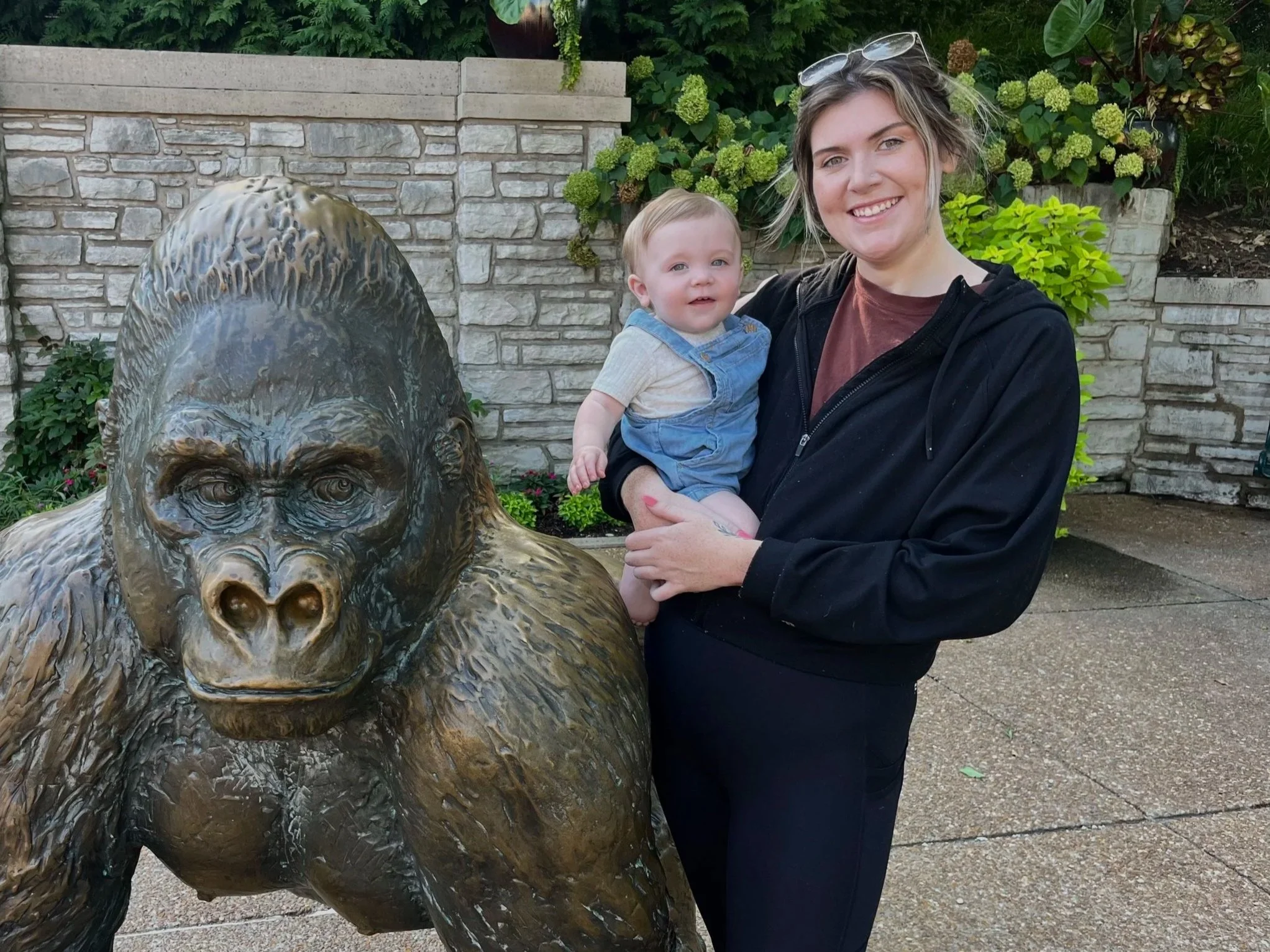 A woman holding a young child standing next to a large bronze statue of a gorilla in a garden setting with plants and a brick wall in the background.