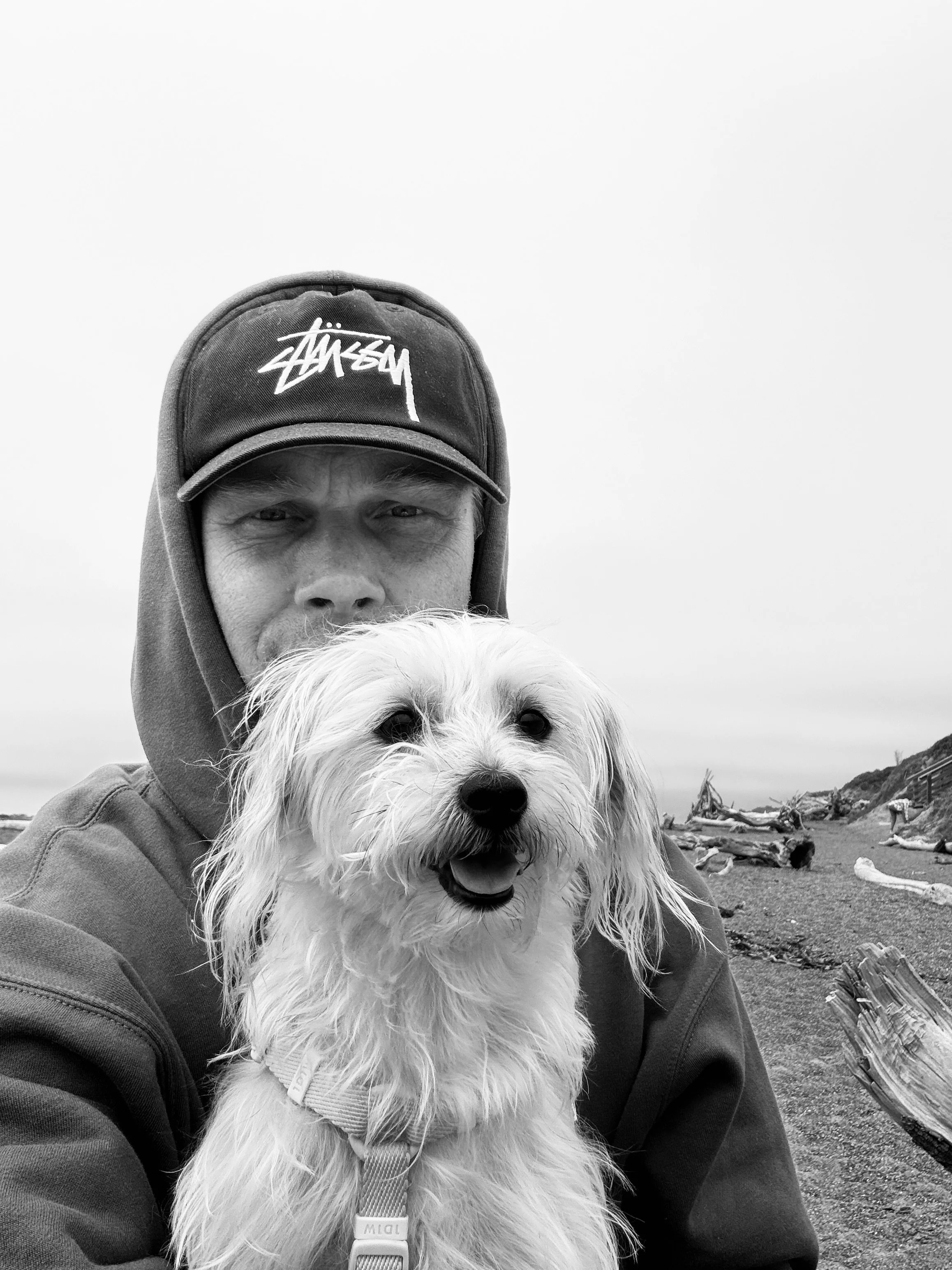 Chris wearing a hoodie and a baseball cap with a logo, holding a small fluffy white dog with a harness, outdoors with driftwood and rocks in the background.