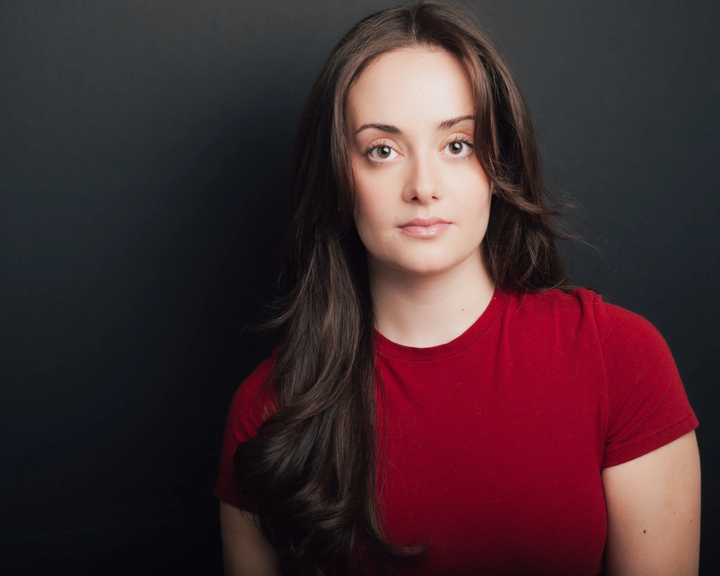 Close-up of a young woman with long brown hair and hazel eyes, wearing a red shirt, against a dark background.