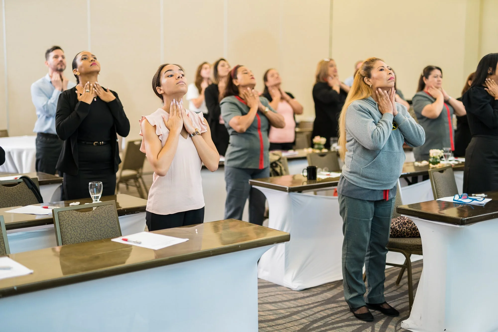 Variedad de participantes en una conferencia o taller, de pie con manos en el cuello o en la garganta, realizando ejercicio de respiración o relajación, en un salón. Hay mesas con papeles, penes y vasos en primer plano.