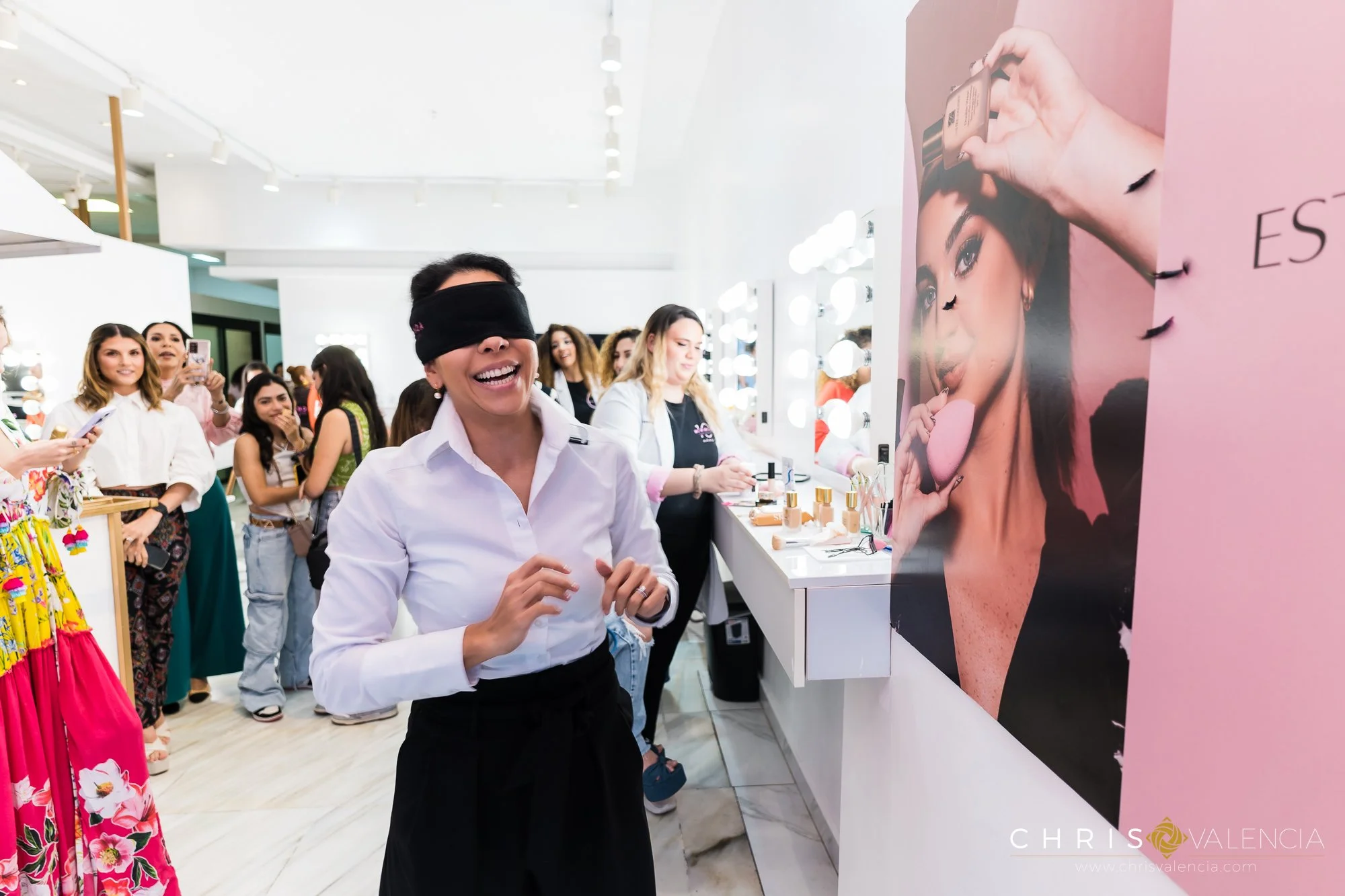 Una mujer con antifaz en un evento de belleza sonríe mientras otras personas la observan y toman fotos en un salón decorado con un cartel grande de una mujer y artículos de maquillaje.