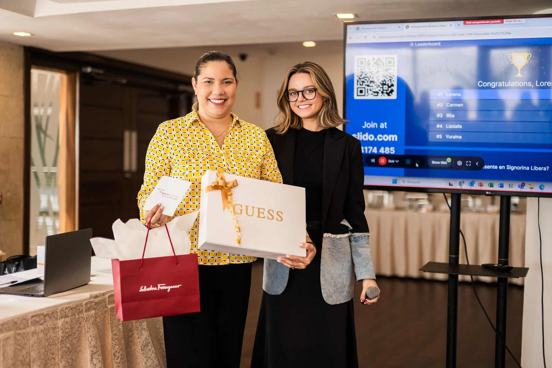Mujeres en un evento de premios o reconocimiento, una de ellas tiene una bolsa de regalo de la marca Salvatore Ferragamo y una caja de Guess. Están sonriendo y posando para la foto, hay una pantalla grande con una lista de ganadores en el fondo.