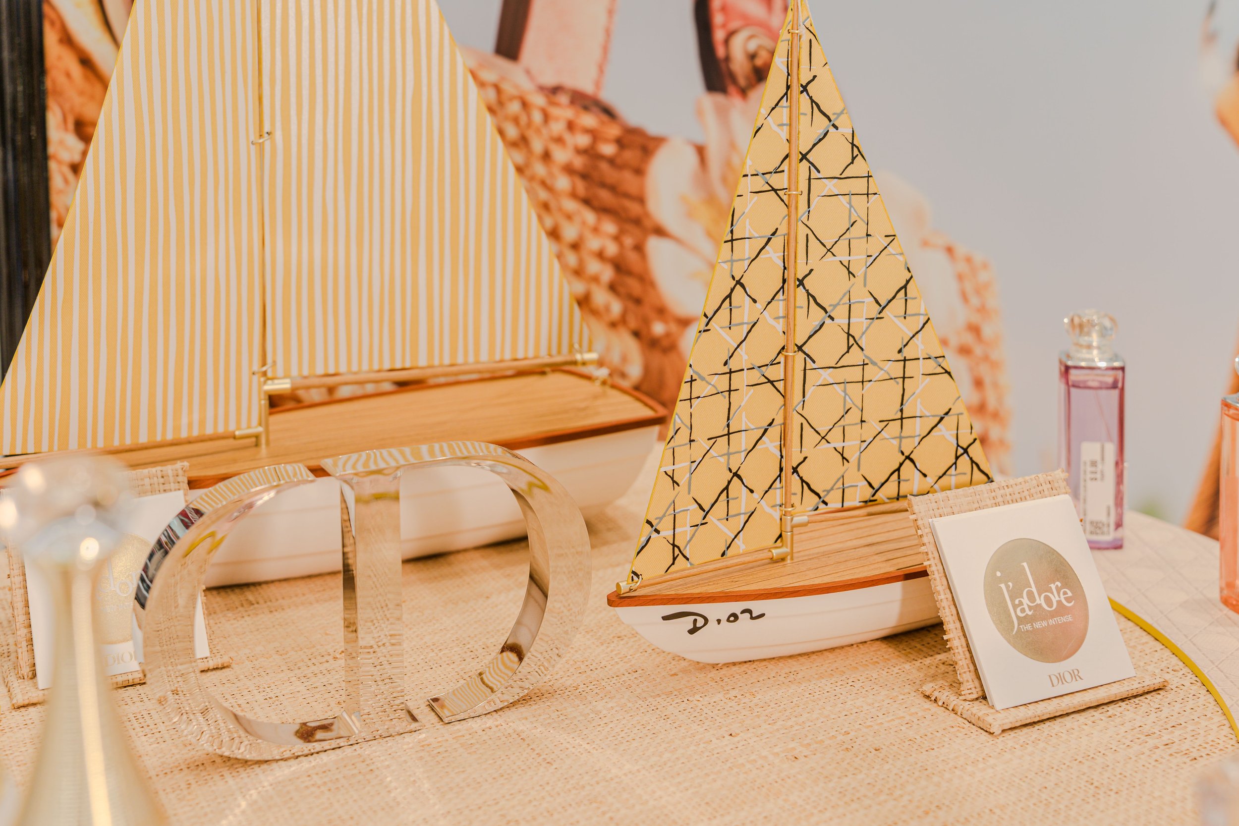 Muestra artículos de belleza y accesorios en una mesa de exhibición, incluyendo un perfume, una tarjeta con la marca Dior y decoraciones en tonos amarillos y beige.