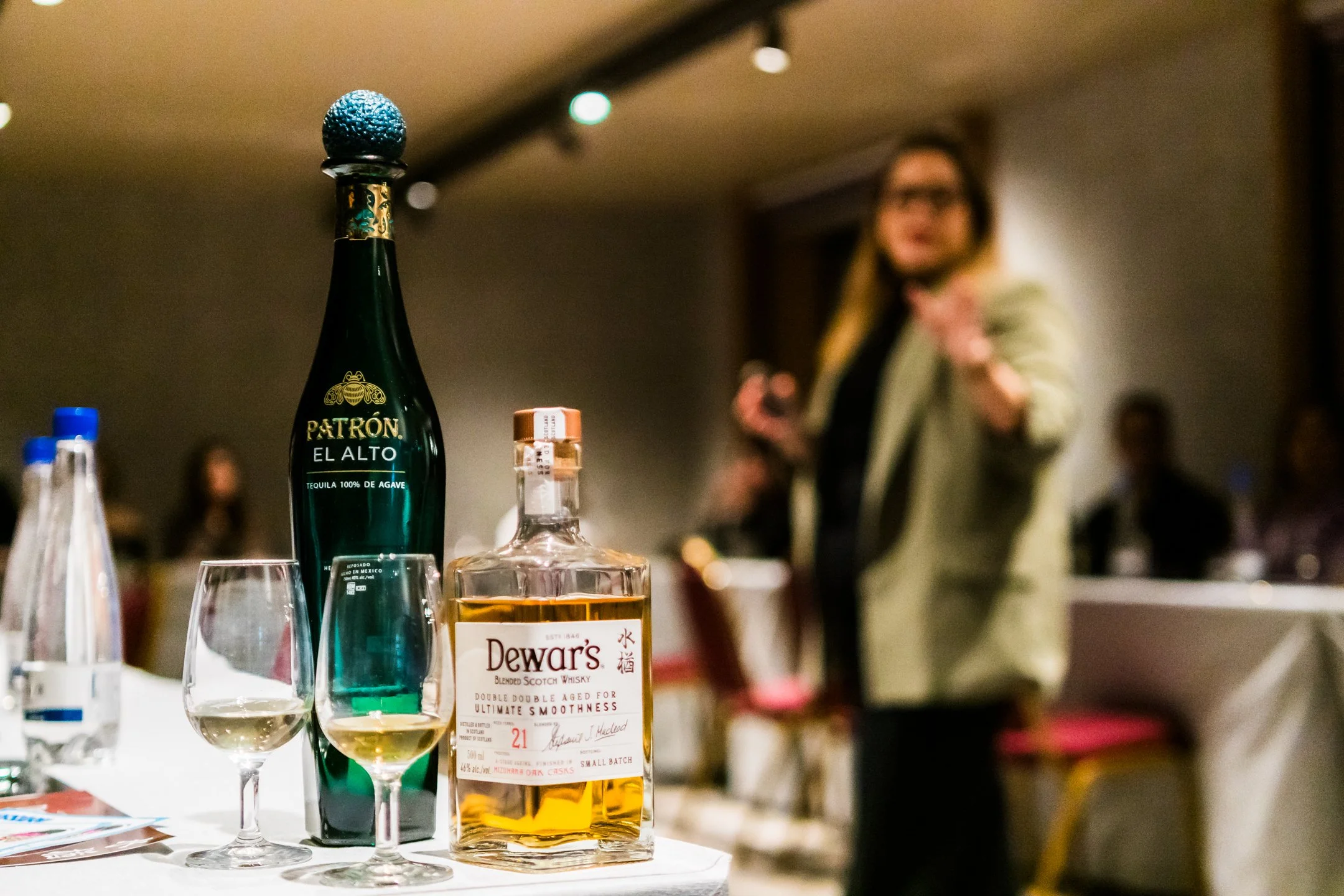 Botellas de licor, una copa con vino, y una botella de whisky en primer plano, en un ambiente de bar con una persona borrosa en el fondo.