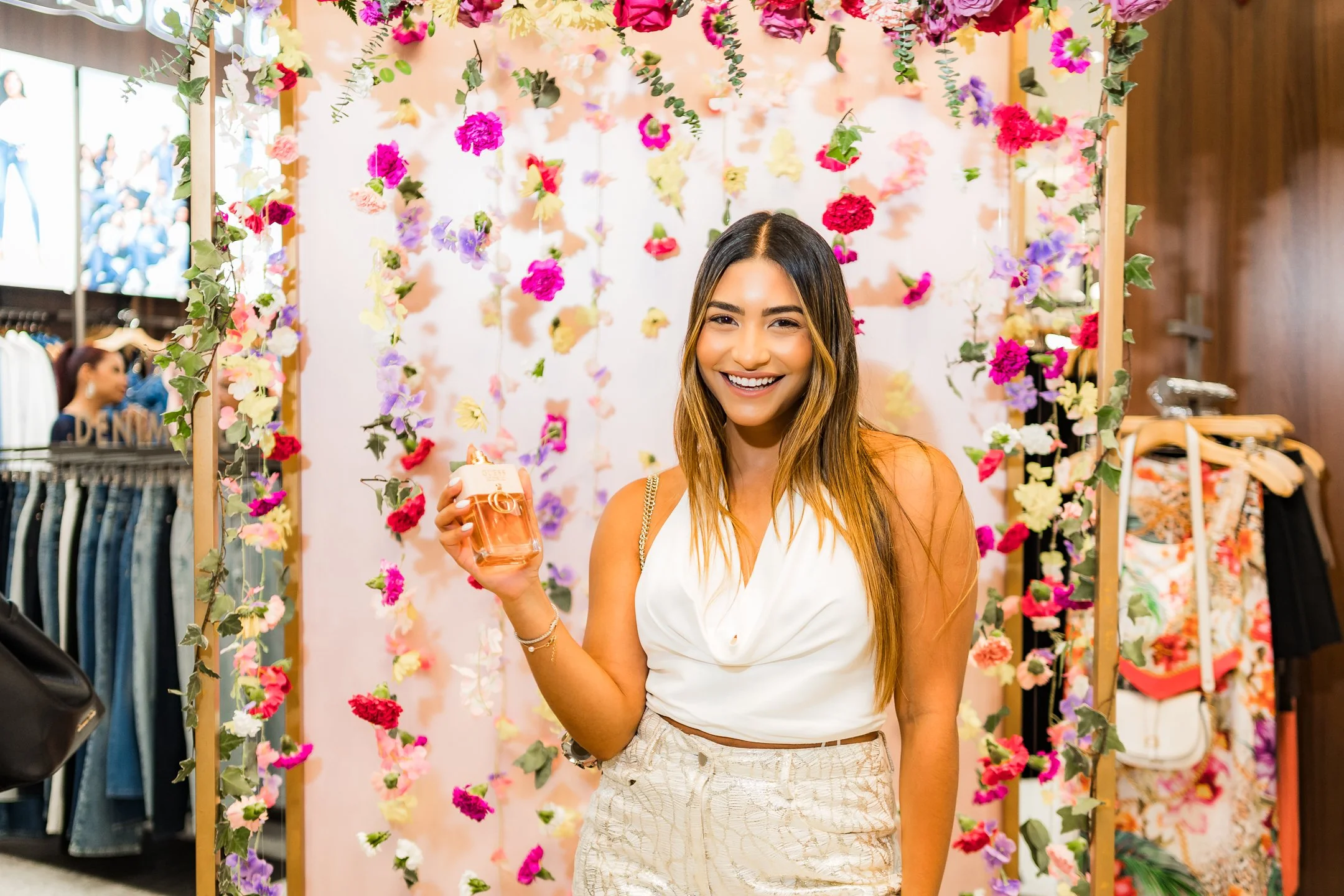 Mujer sonriendo sosteniendo una taza de bebida en un decorado de flores en un entorno de tienda de ropa.