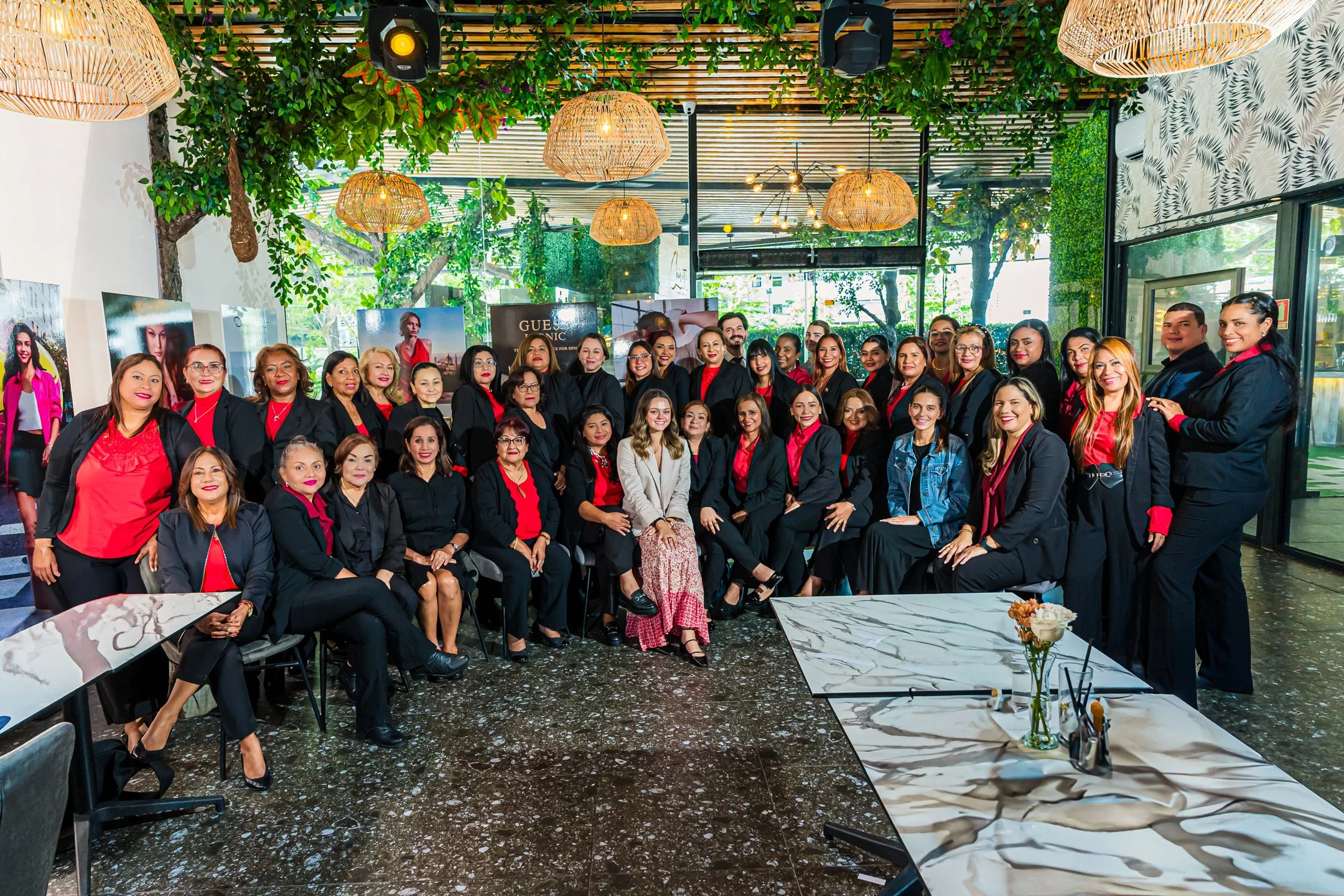 Grupo de mujeres y algunos hombres posando para una foto en un lugar decorado con plantas y lámparas de mimbre, algunas con ropa formal en tonos negros y rojos, en un ambiente con paredes decoradas con papel tapiz de hojas y ventanas grandes con vist