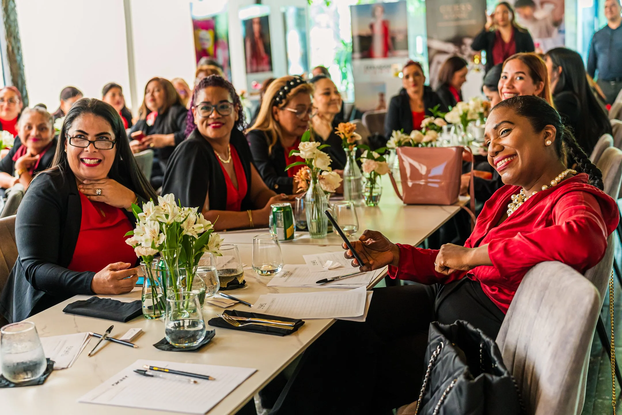 Un grupo de mujeres sentadas en una mesa en una reunión o evento social, algunas sonriendo y otras usando dispositivos electrónicos. La mesa tiene flores y utensilios, y la sala tiene un ambiente luminoso y colorido.