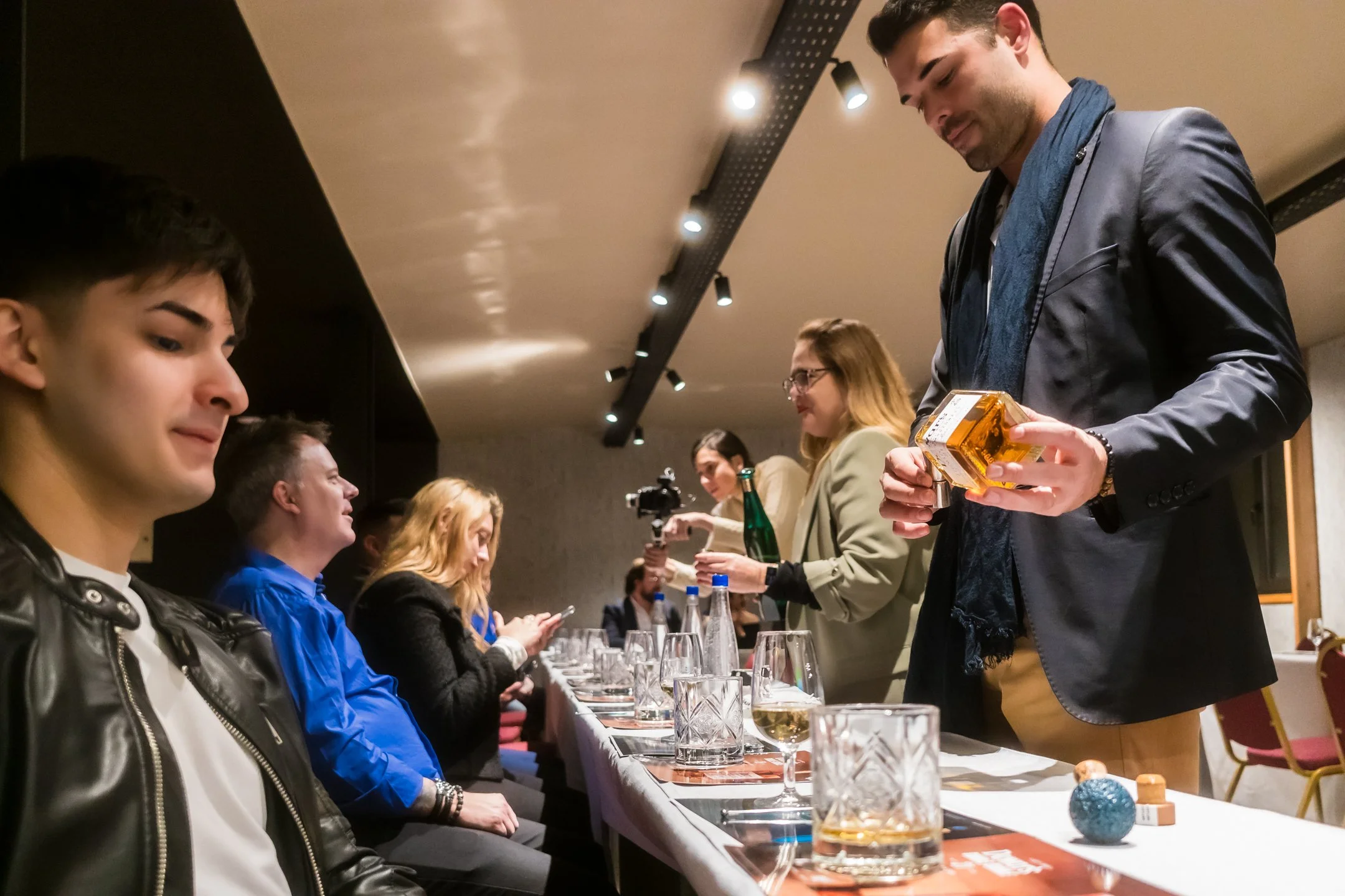 Grupo de personas en una cena, con un camarero entregando una botella de vino a una mujer, en un ambiente interior con iluminación puntual.