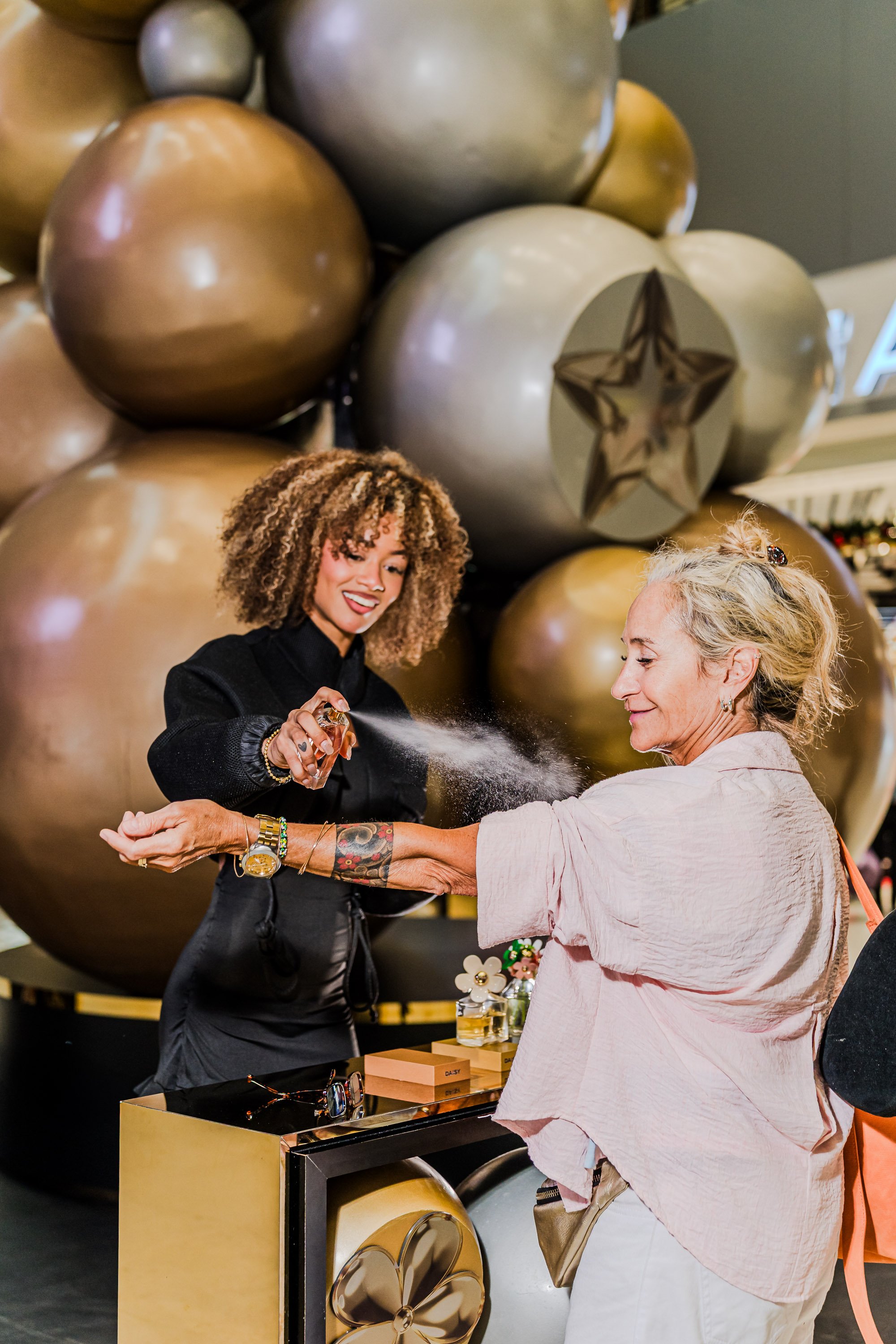 Mujer con cabello rizado y mujer de cabello rubio, ambas sonriendo y mostrando accesorios de gafas y pulseras, en una tienda de accesorios, con globos dorados y plateados en el fondo.