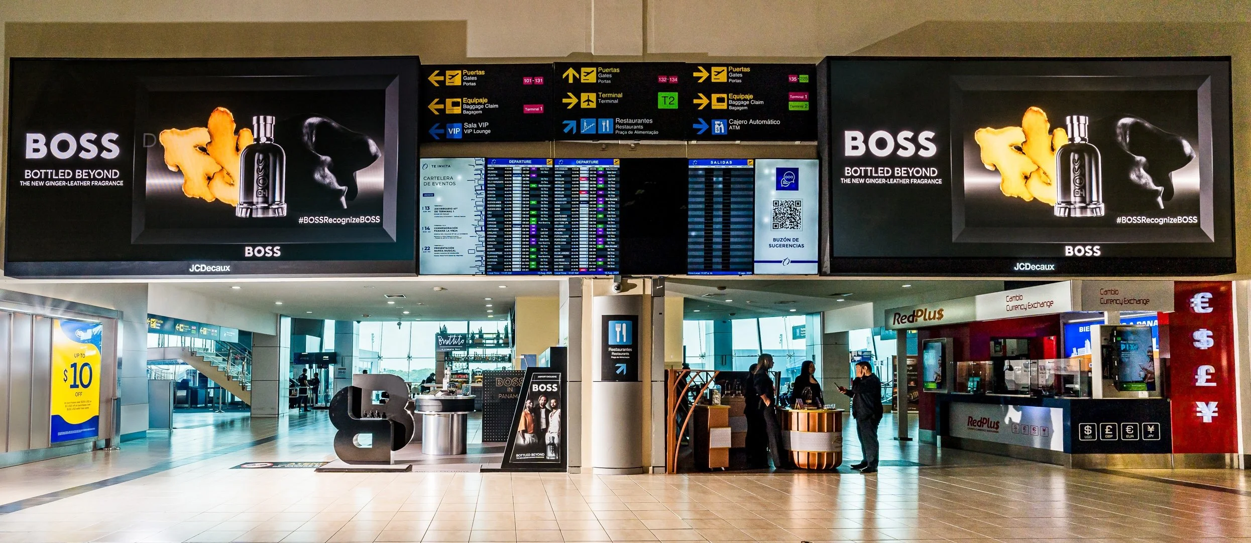 Pantalla de informaciones y relojes de vuelos en un aeropuerto, con publicidad de perfume Boss y personas en el fondo en el área de salidas y llegadas.