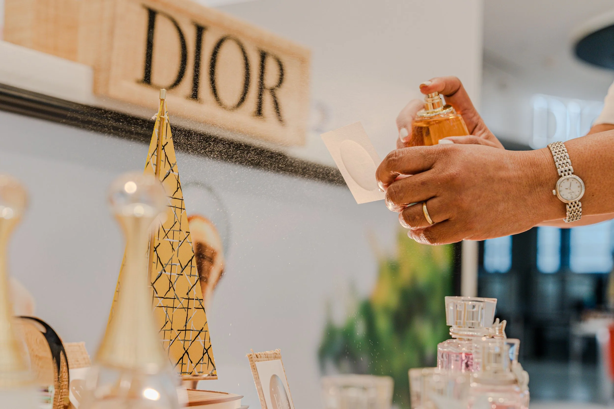 Persona rociando perfume en una mano, con exhibición de artículos de belleza y decoraciones en el fondo.