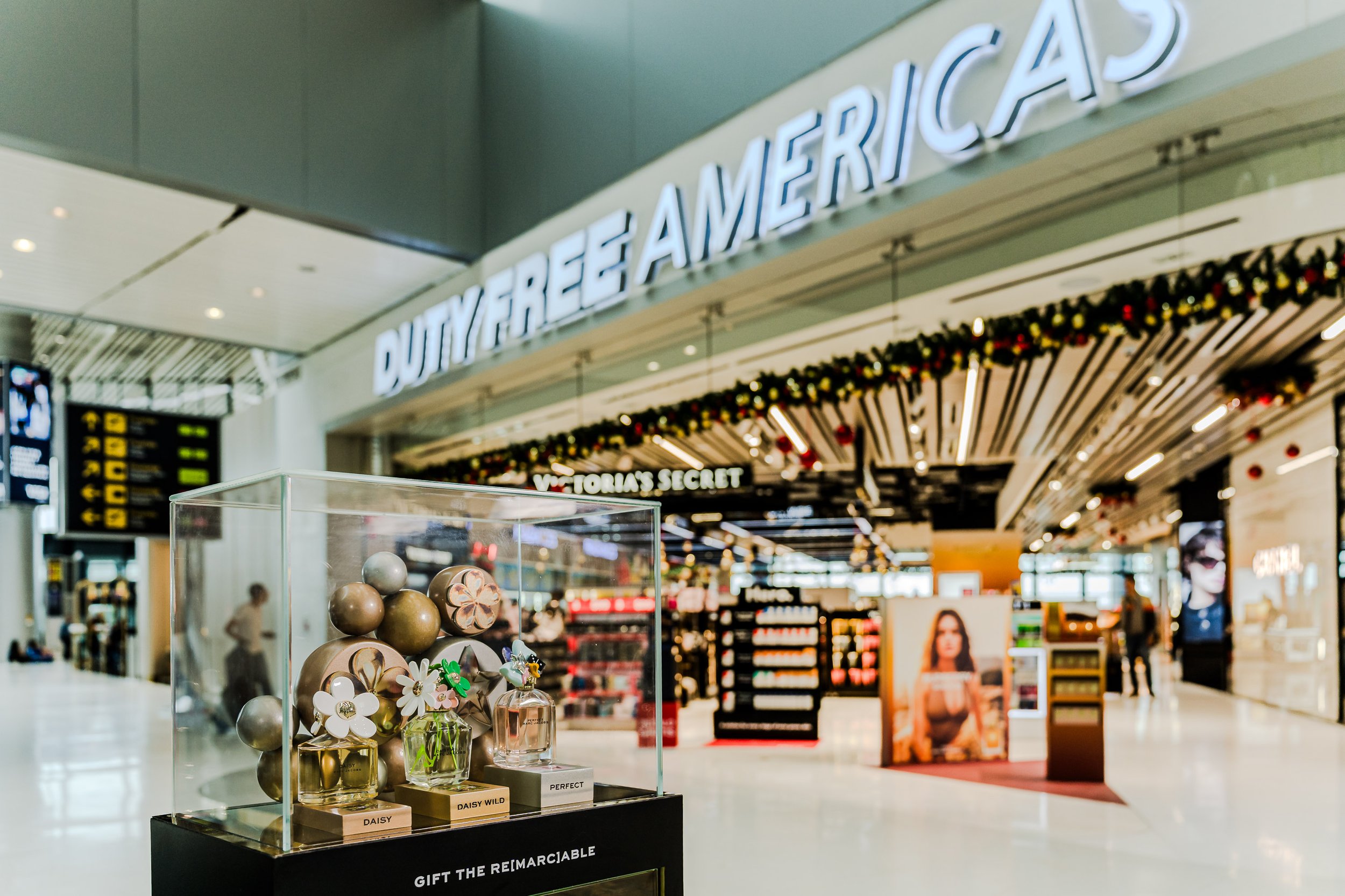 Exhibición de perfumes en una tienda de un aeropuerto, con el letrero 'Duty Free America' visible en el fondo y decoraciones navideñas.