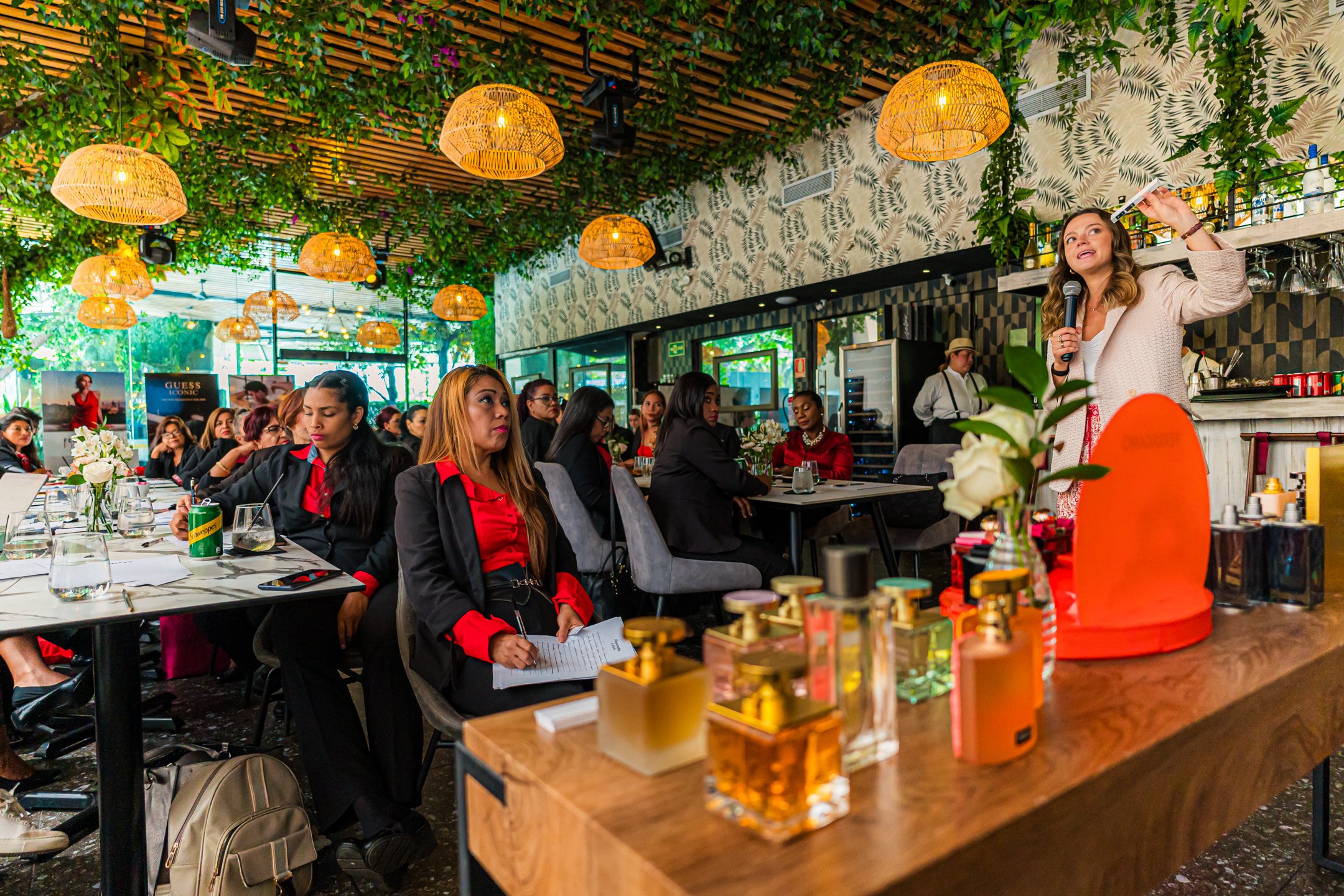 Mujer dando una charla con micrófono en un restaurante o evento, con público atento, decoraciones con plantas y lámparas de mimbre, en un ambiente moderno y colorido.