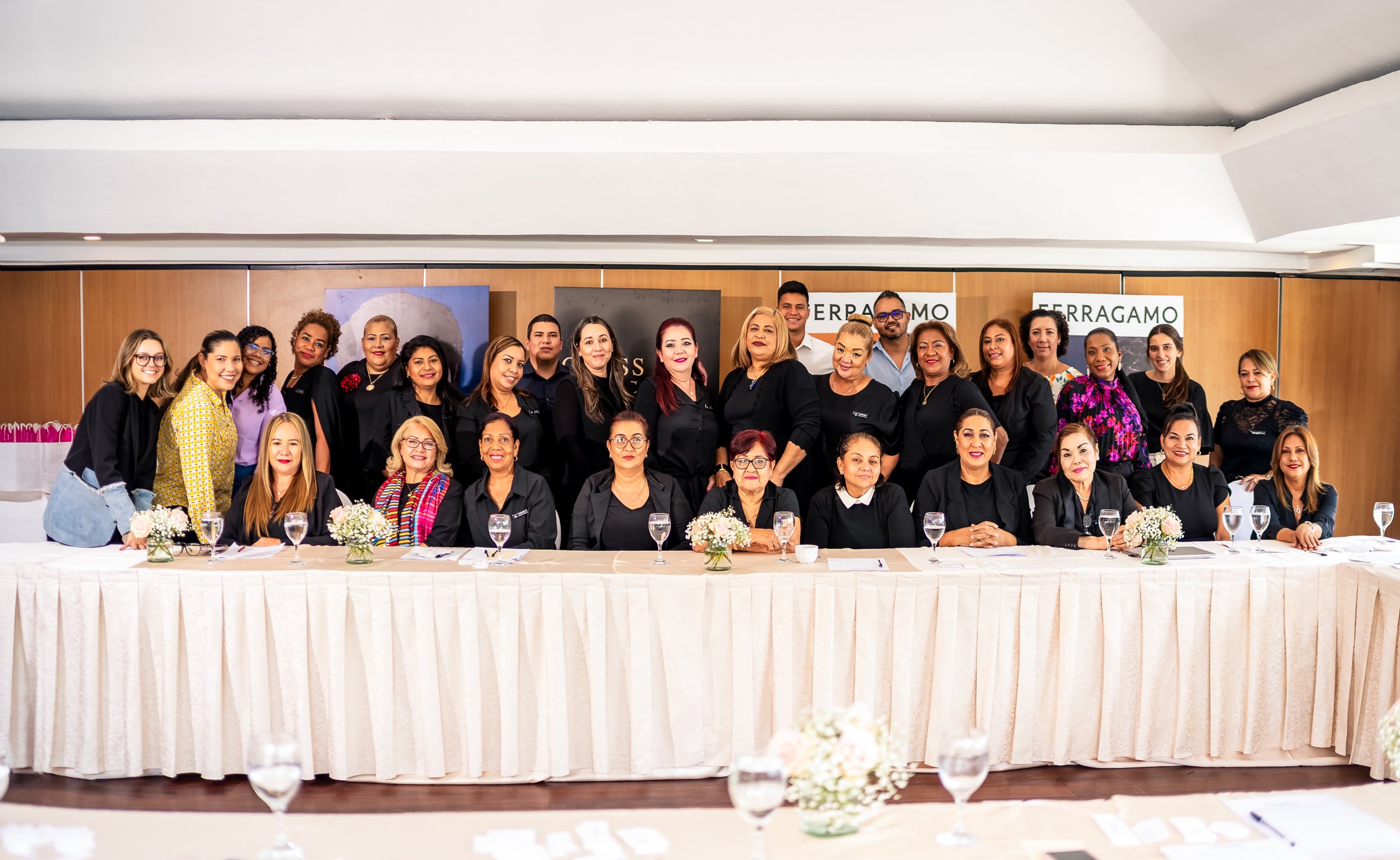 Grupo de mujeres y algunos hombres posando en una reunión o evento formal, frente a un fondo con carteles que dicen 'Ferragamo'. Están sentados y de pie tras una mesa con flores y vasos de agua.