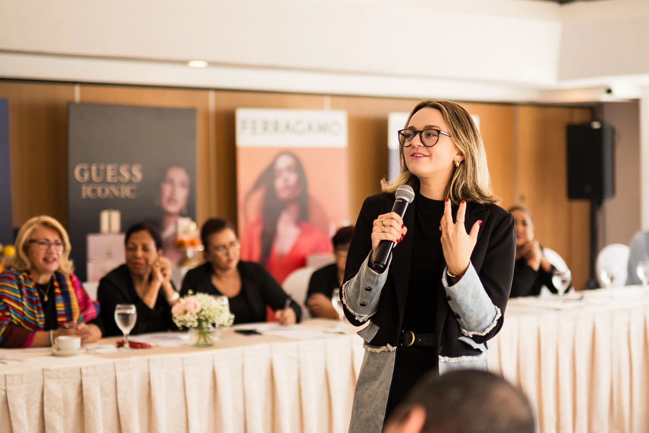 Mujer con gafas y blazer hablando con micrófono en una reunión con varias mujeres en la mesa, en un salón con carteles de moda al fondo.