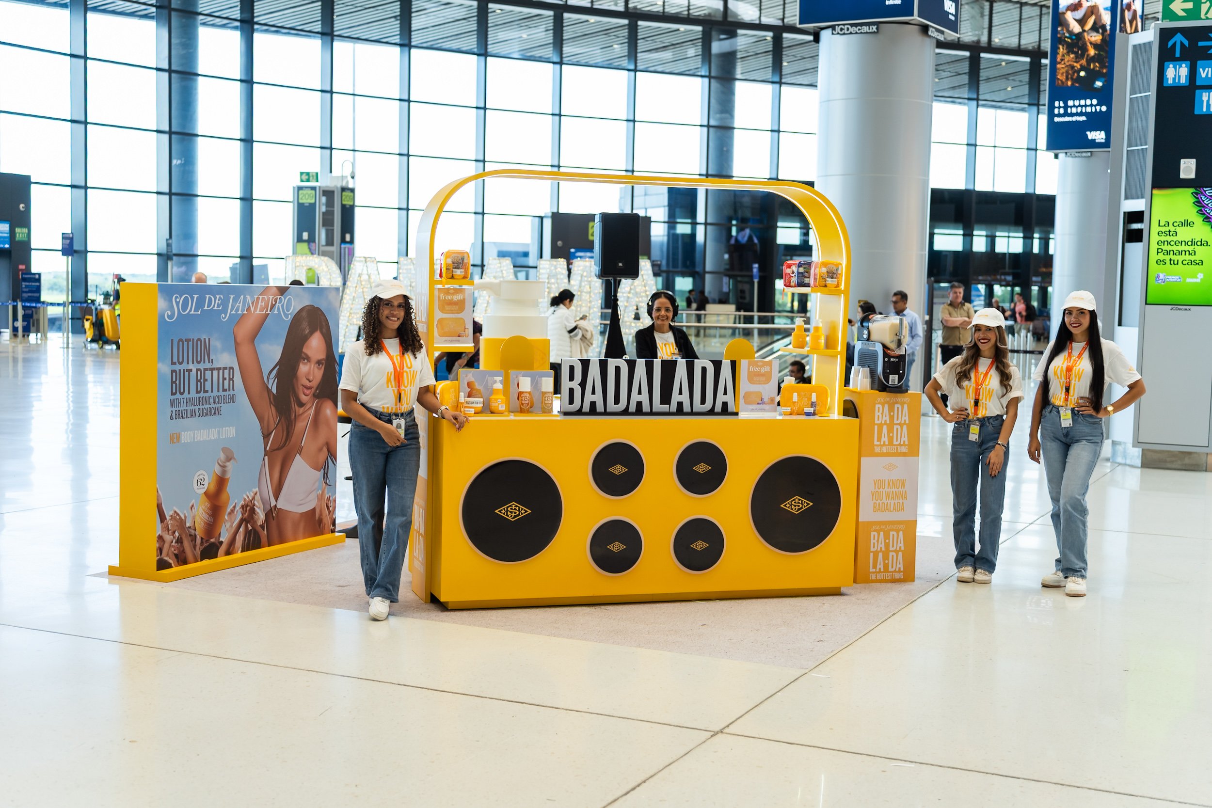 Mostrador en aeropuerto promocionando productos de cuidado de la piel con un cartel que dice 'Sol de Janeiro' y una mujer en un cartel publicitario. El mostrador es de color amarillo y hay tres mujeres sonriendo detrás de él, con ropa blanca y gorros