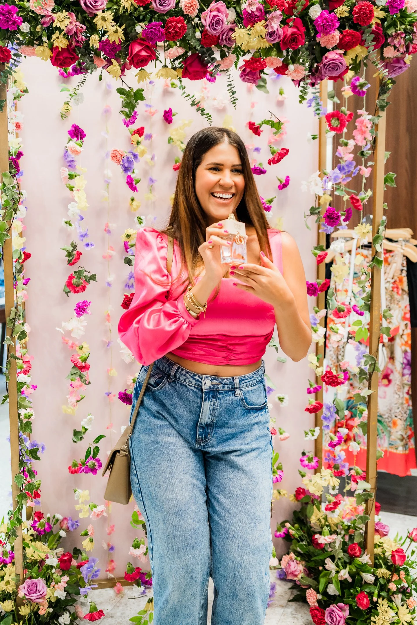 Mujer sonriendo con vestido rosa y jeans, rodeada de decoraciones florales coloridas en un fondo blanco en un entorno interior.