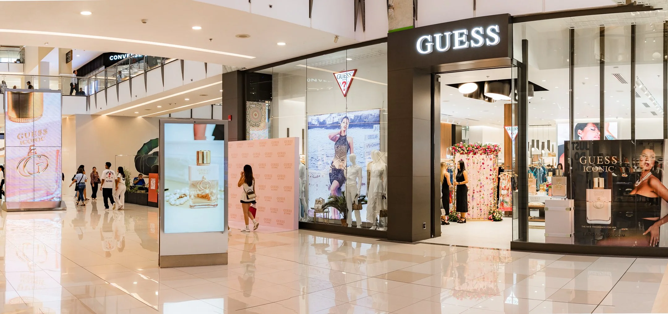 Entrada a la tienda de moda Guess en un centro comercial, con varias pantallas digitales y personas caminando y comprando.