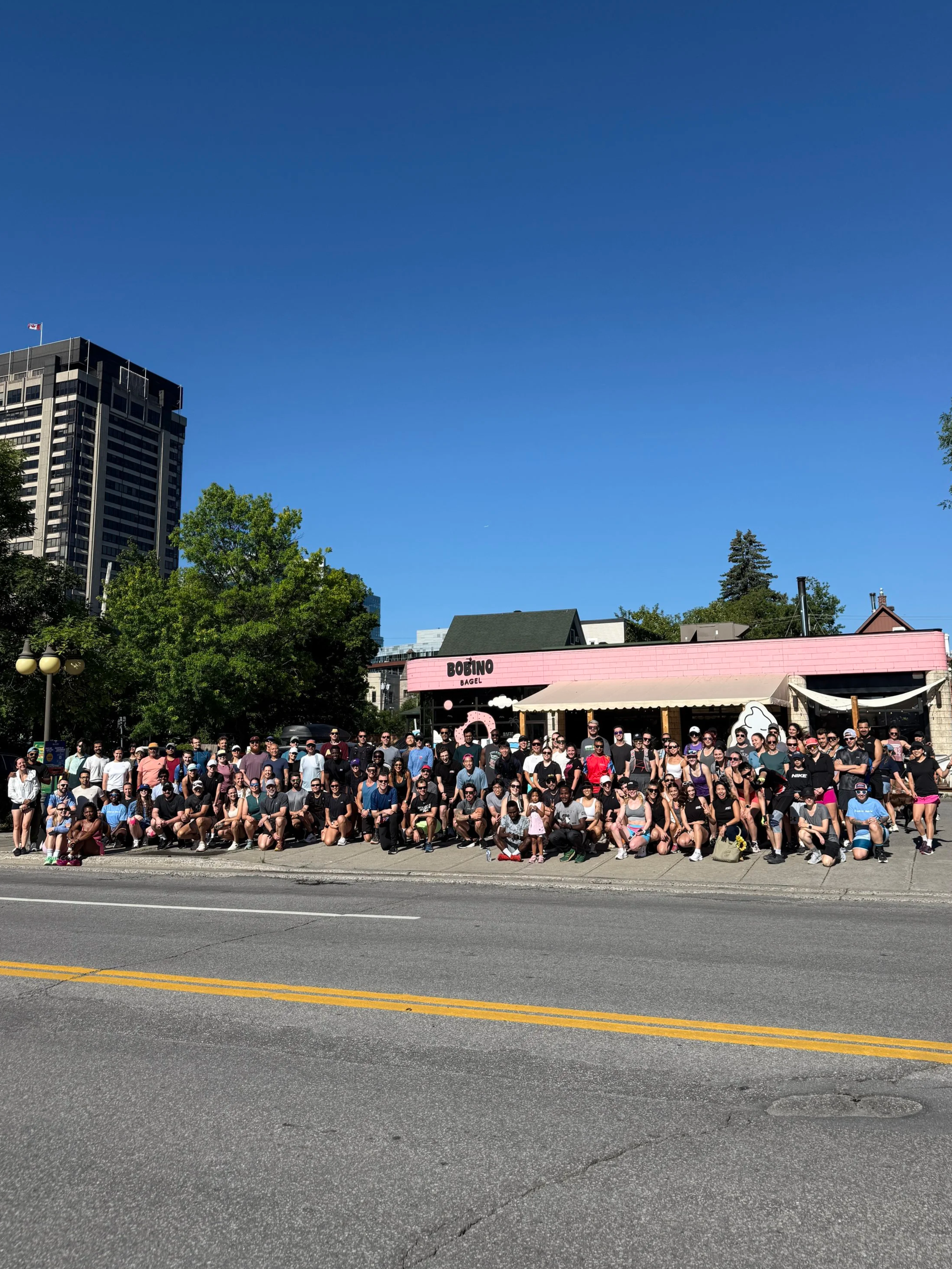 Large group of people gathered outside in front of a pink building with sign 'Bobino Bagel'. The group includes adults and children, all standing or sitting on the sidewalk. It appears to be a sunny day with clear blue sky and some trees and high-rise buildings in the background.