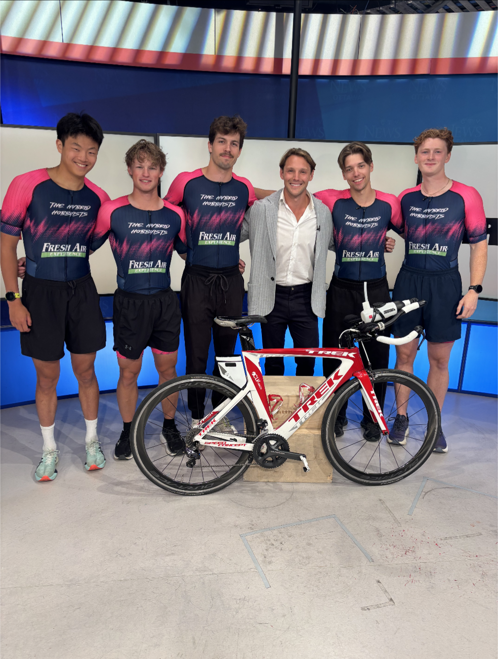 Six young men in matching blue and pink cycling jerseys stand behind a red and white Trek racing bicycle, with one man in a gray blazer and white shirt in the center, in a TV studio.