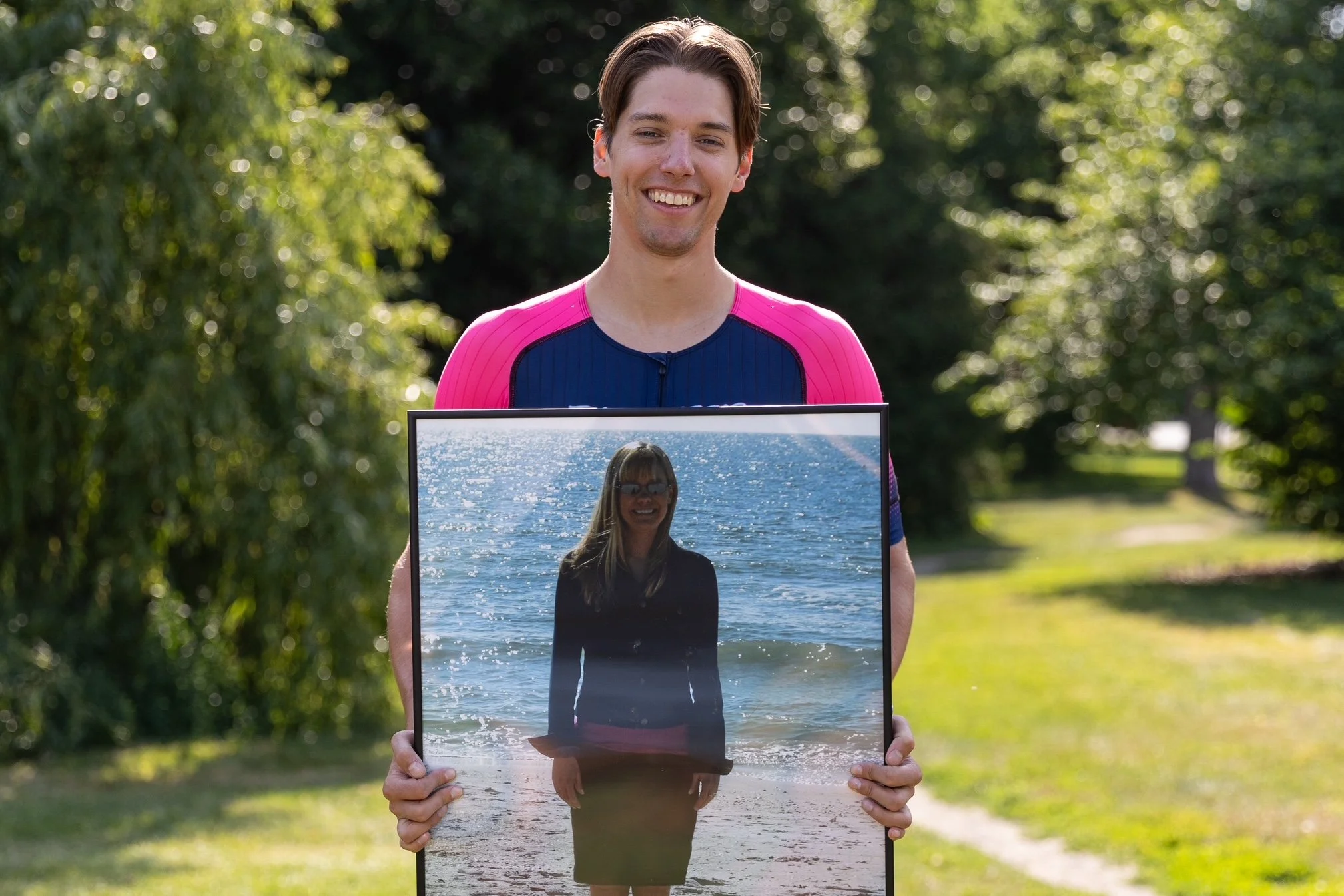 A smiling young man in a blue and pink athletic shirt holds a framed image of a woman at the beach, with green trees in the background.