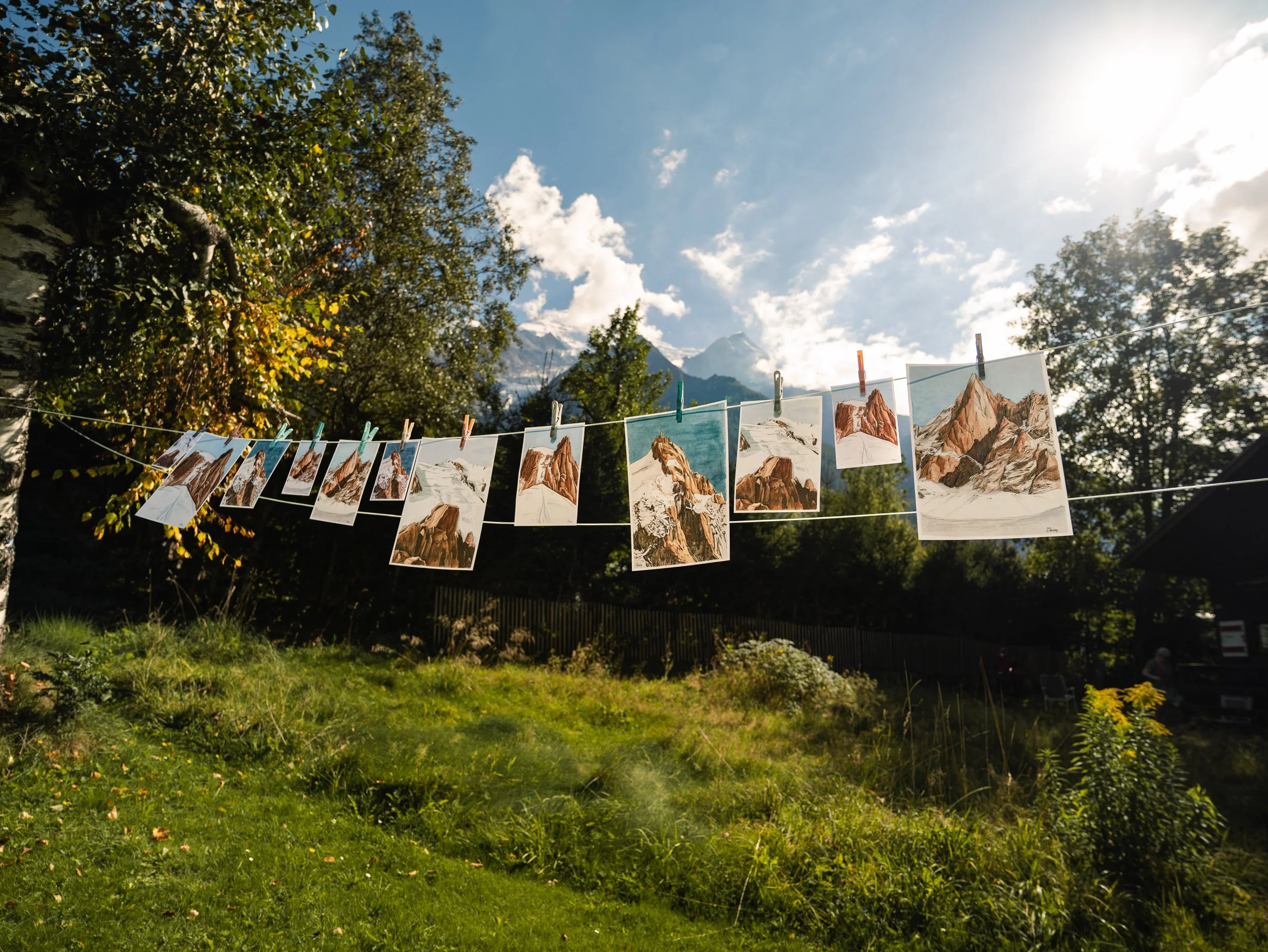 Photographie de vêtements ou de dessins de paysages de montagne suspendus sur une corde à linge en plein air, avec arbres et montagnes en arrière-plan sous un ciel partiellement nuageux, ensoleillé.