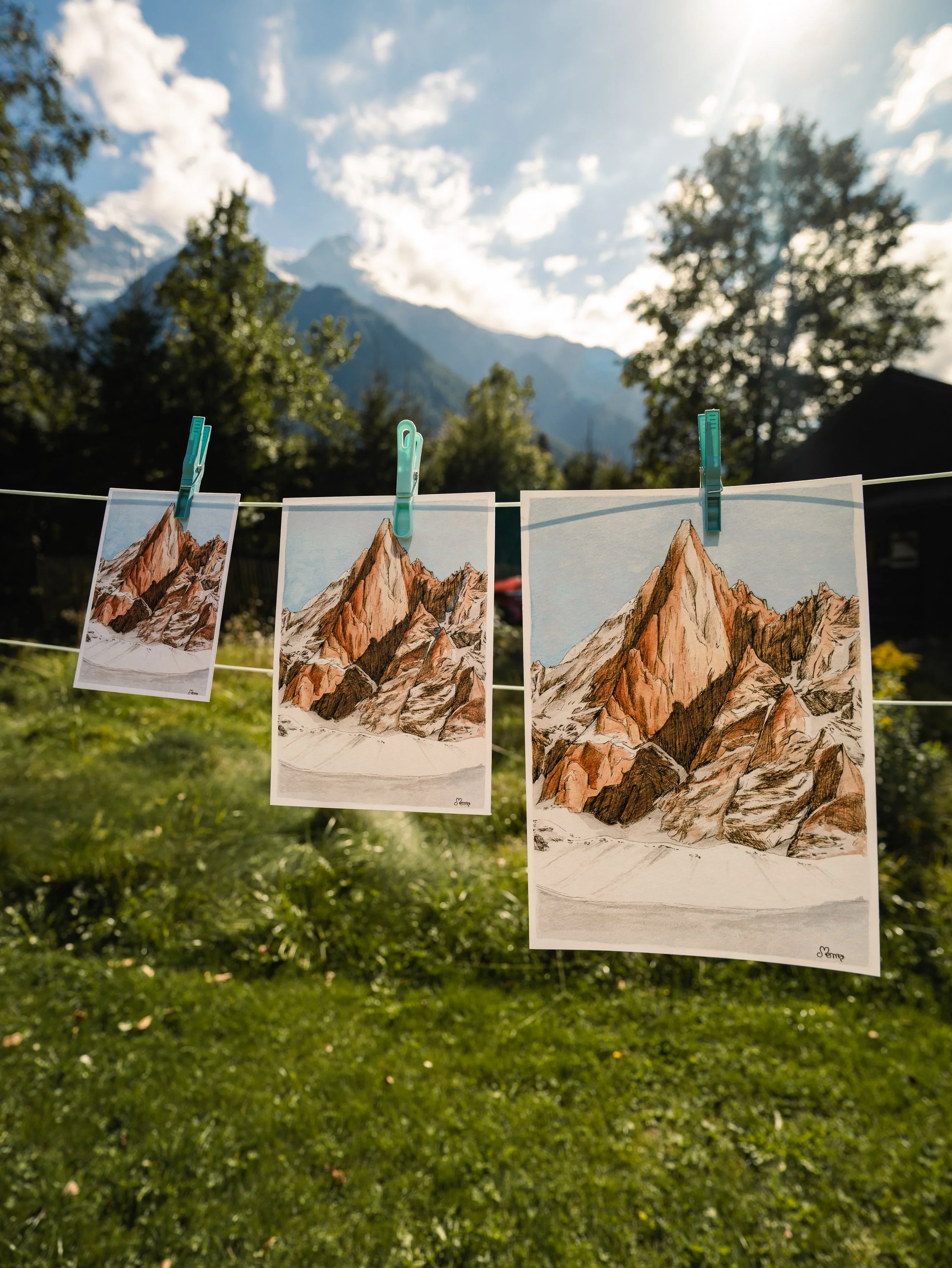 Trois dessins de montagnes accrochés à une corde avec des pinces à linge, en extérieur avec un ciel bleu et des montagnes en arrière-plan.