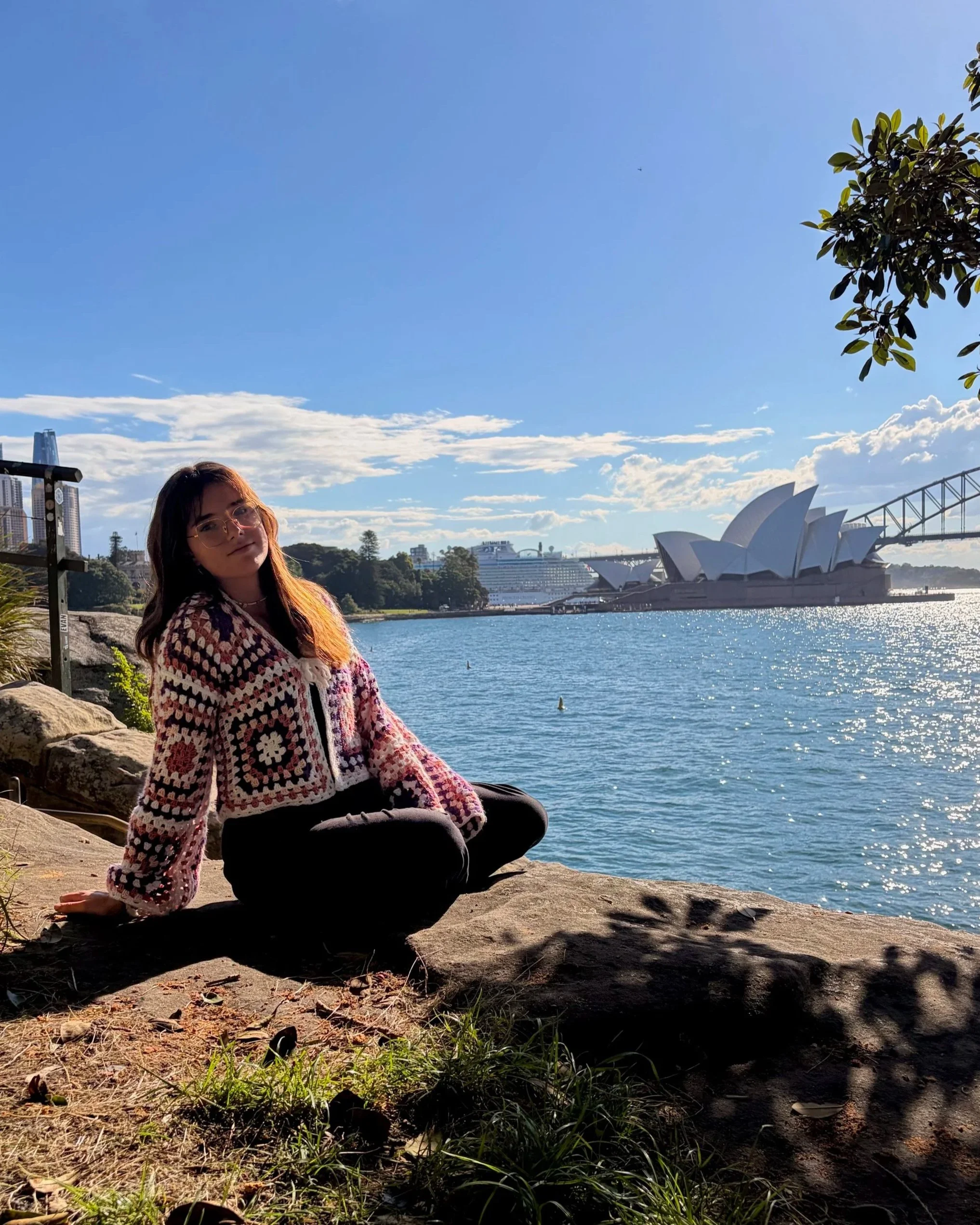 Une femme portant un pull à motifs colorés est assise sur un rocher près d'un plan d'eau, avec la ville de Sydney, dont l'Opéra, en arrière-plan par une journée ensoleillée.