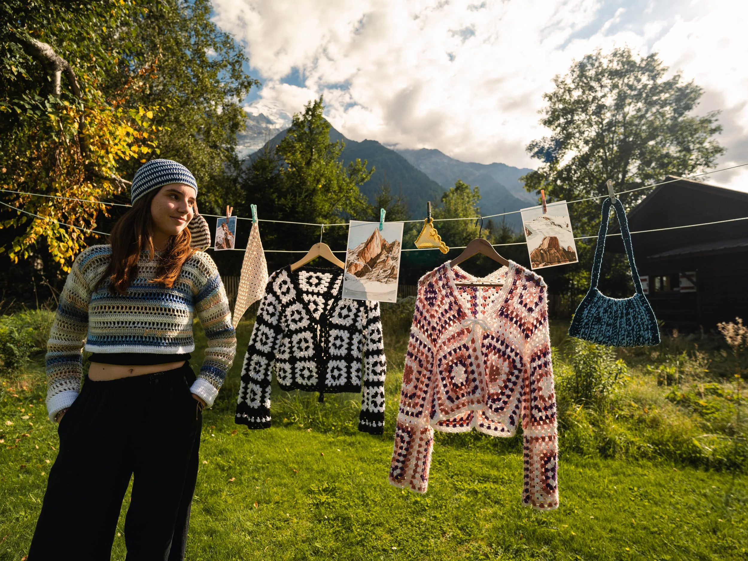 Une jeune femme avec un pull en crochet rayé et un bonnet regarde une corde à linge où sont suspendues des vêtements en crochet et des photos.