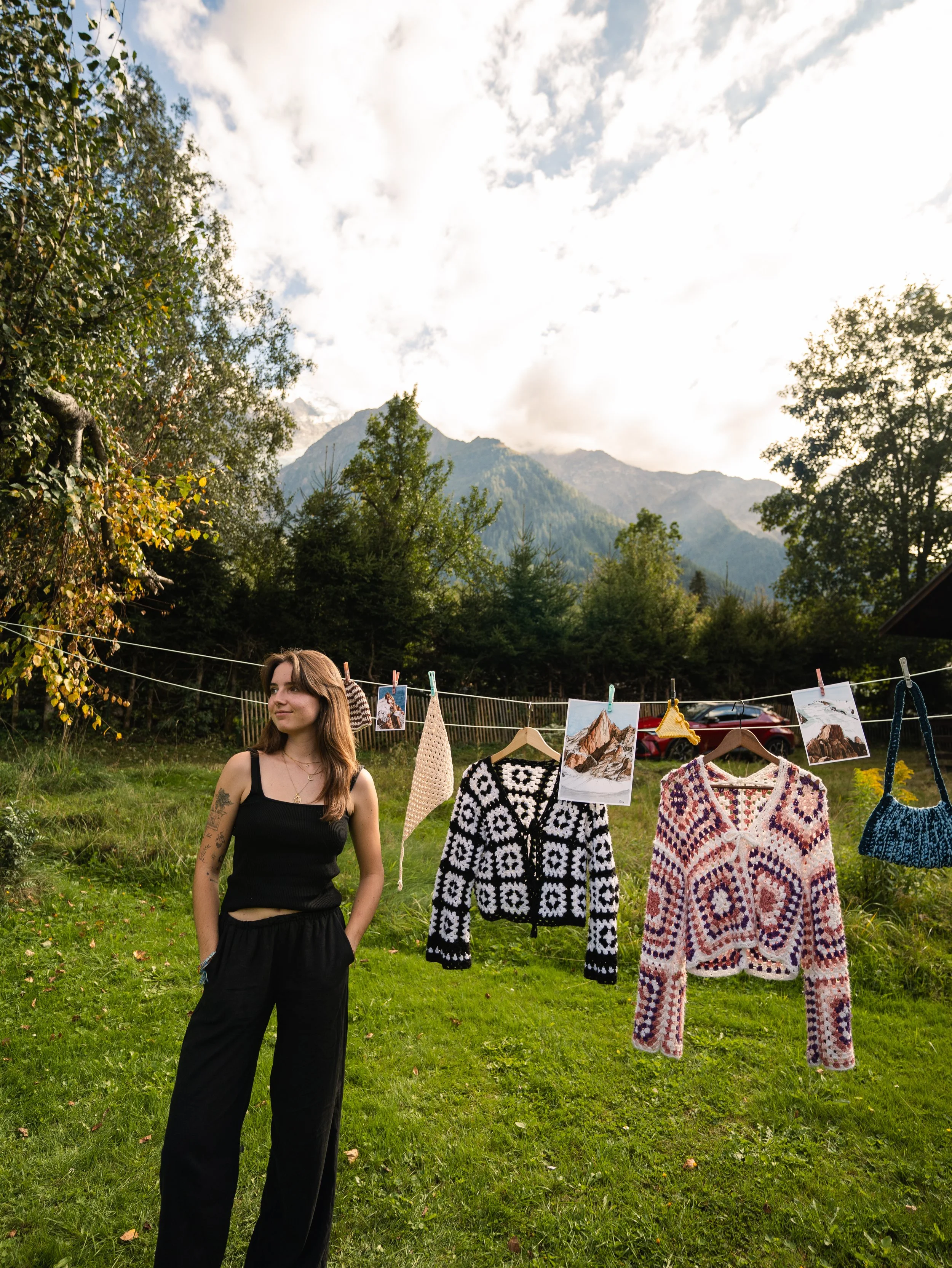 Une jeune femme avec des cheveux longs et des tatouages sur le bras, vêtue d'une robe noire, se tenant dans un champ d'herbe verte, avec une cordeline à linge où sont suspendues des photos et des vêtements tricotés, un arbre et des montagnes en arrière-plan sous un ciel partiellement nuageux.