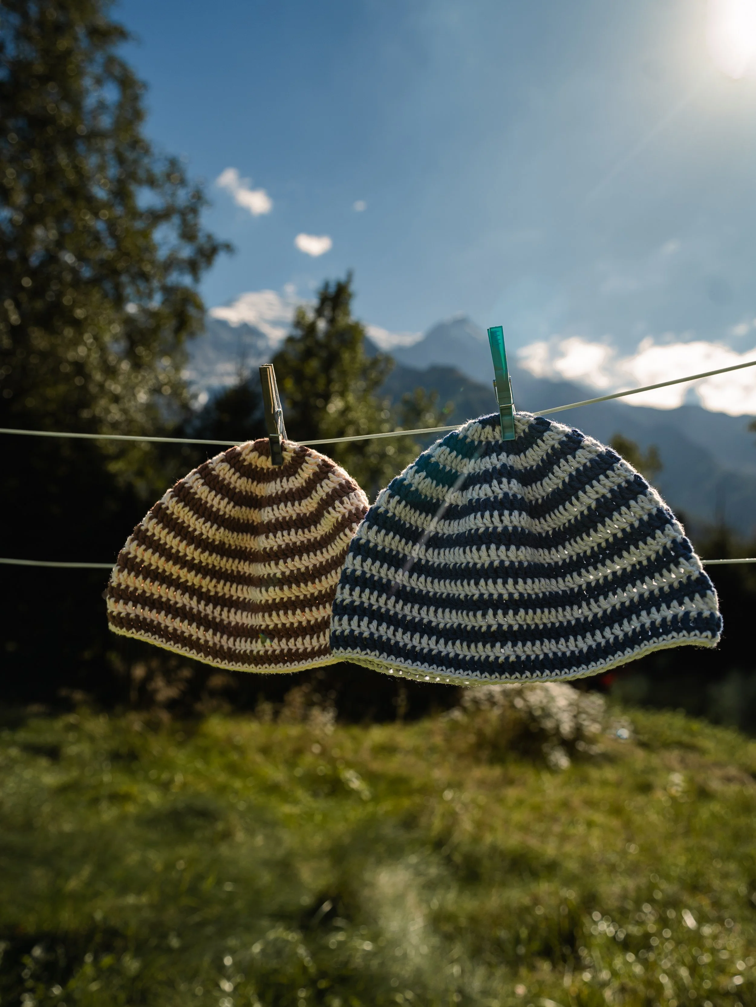 Deux bonnets tricotés suspendus sur une corde à linge avec des pinces, en plein air avec un ciel bleu et des montagnes en arrière-plan.