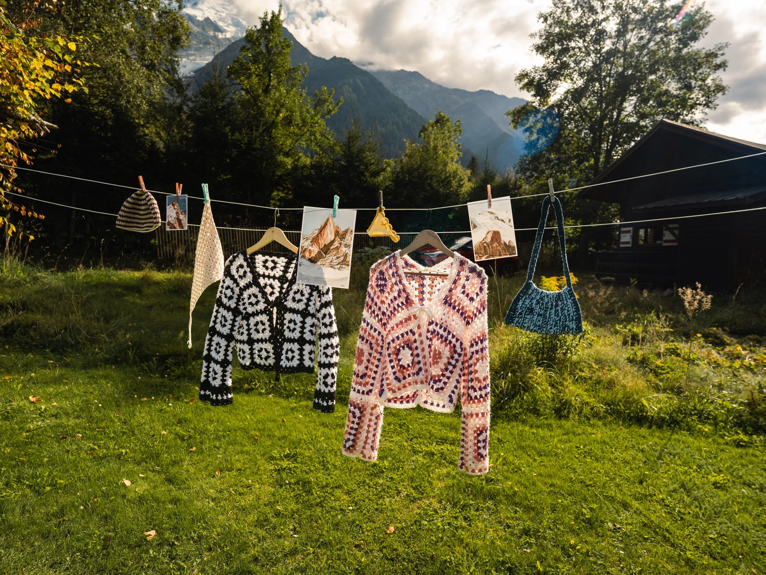 Des vêtements tricotés sont suspendus à une corde à linge en plein air, avec un paysage de montagnes et d'arbres en arrière-plan.