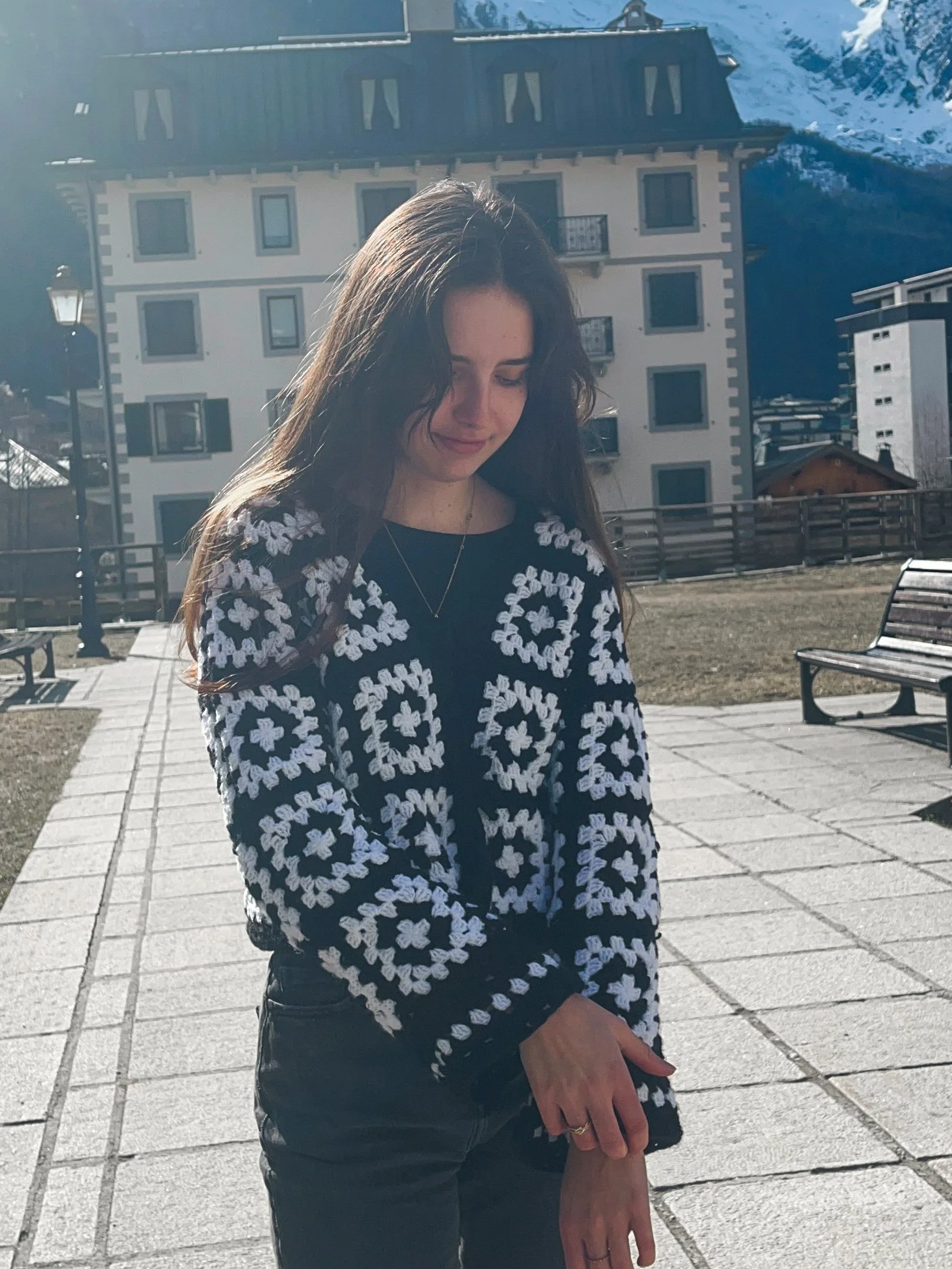 Une jeune femme avec de longs cheveux bruns, portant un pull noir avec un motif de fleurs blanches, se tenant à l'extérieur dans un endroit de style montagne avec des bâtiments en arrière-plan, sous un ciel ensoleillé.