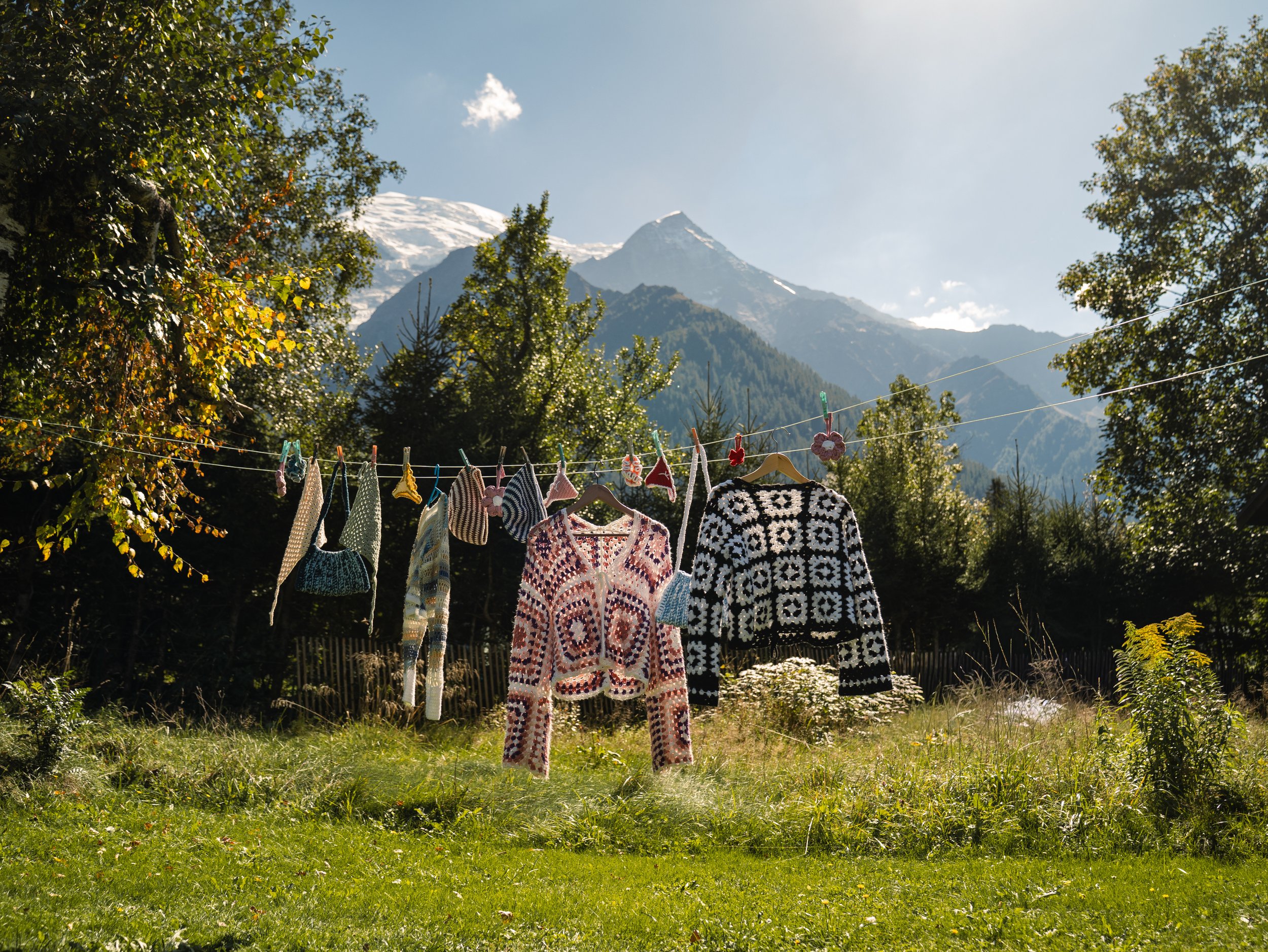 L'image montre une corde à linge avec des vêtements en laine tricotés, suspendus pour sécher, dans un jardin entouré d'arbres et de montagnes enneigées en arrière-plan.