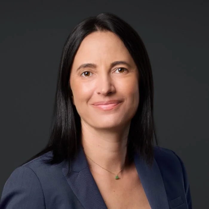 Professional portrait of a woman with shoulder-length black hair, wearing a navy blazer and a delicate necklace, against a dark gray background.