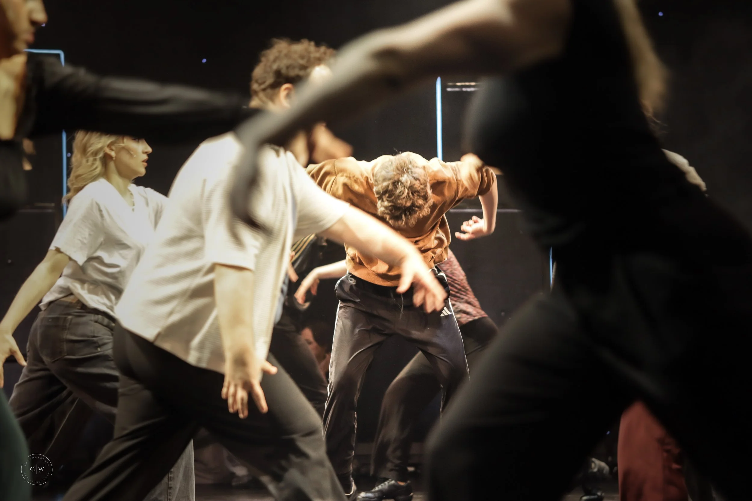 Group of dancers performing on stage in a dark setting, captured mid-movement with focus on their bodies and attire.