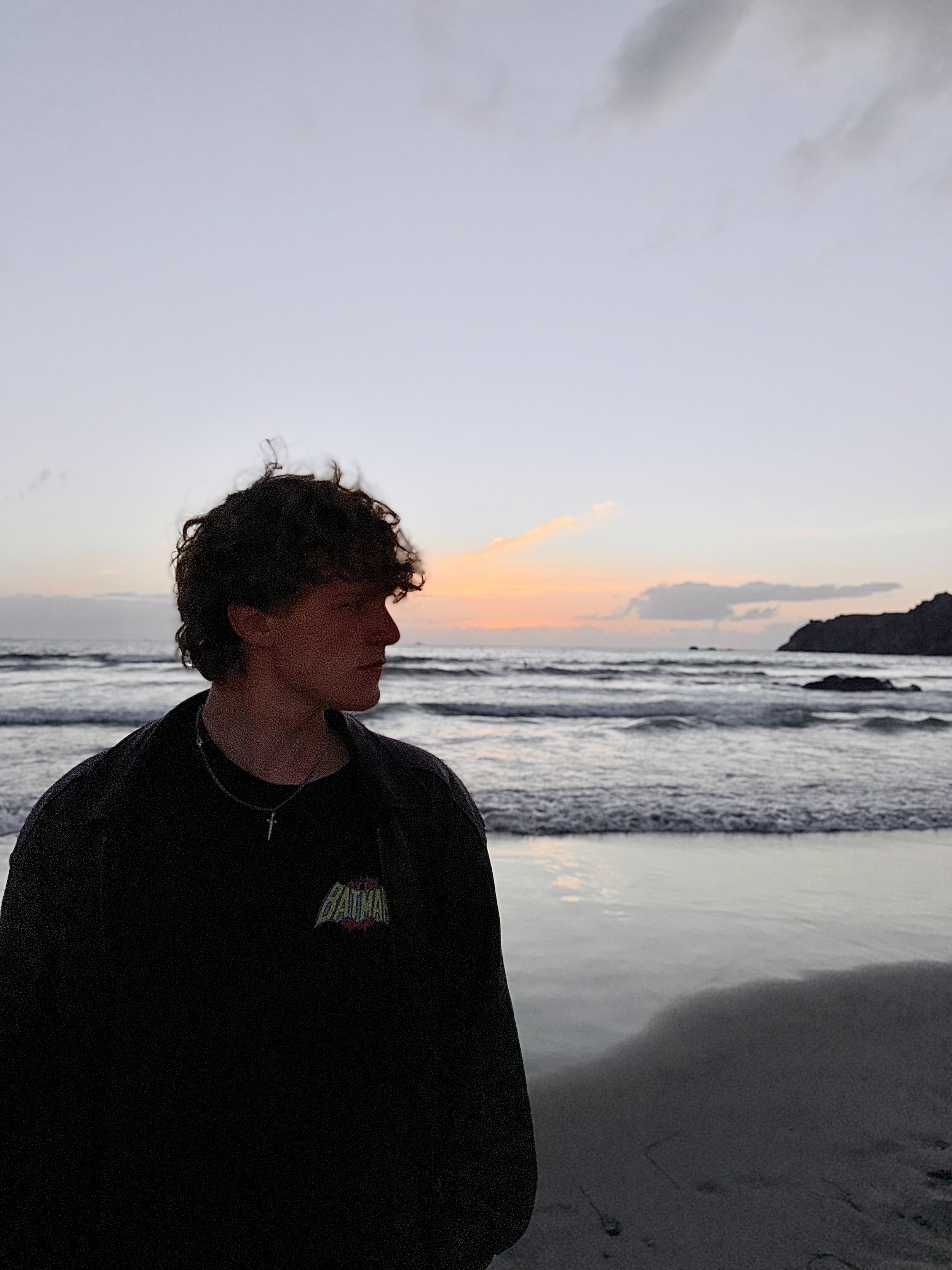 A person with curly hair standing on a beach at sunset, with the ocean and cliffs in the background.