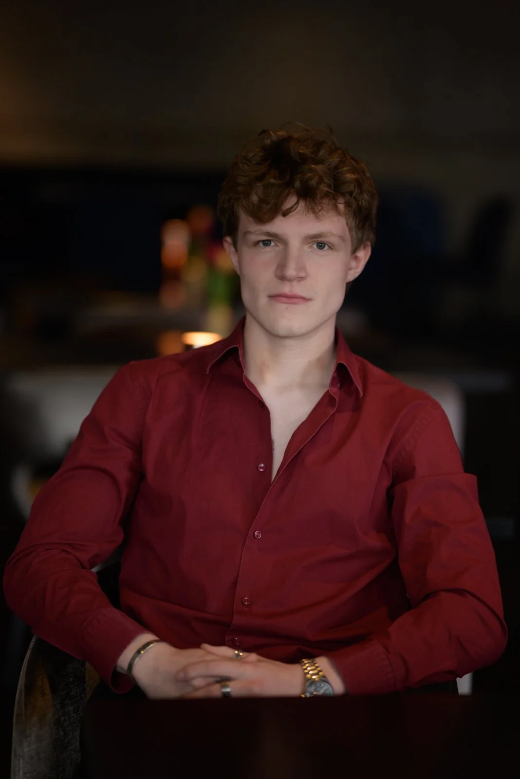A young man with curly red hair and fair skin, wearing a burgundy dress shirt, sits with his hands clasped on a table, looking directly at the camera in a dimly lit room with blurred background.