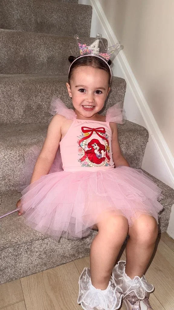 A young girl dressed as a princess, sitting on carpeted stairs. She is wearing a pink tutu dress with Ariel from The Little Mermaid on it, a sparkly crown headband, and fancy shiny shoes.