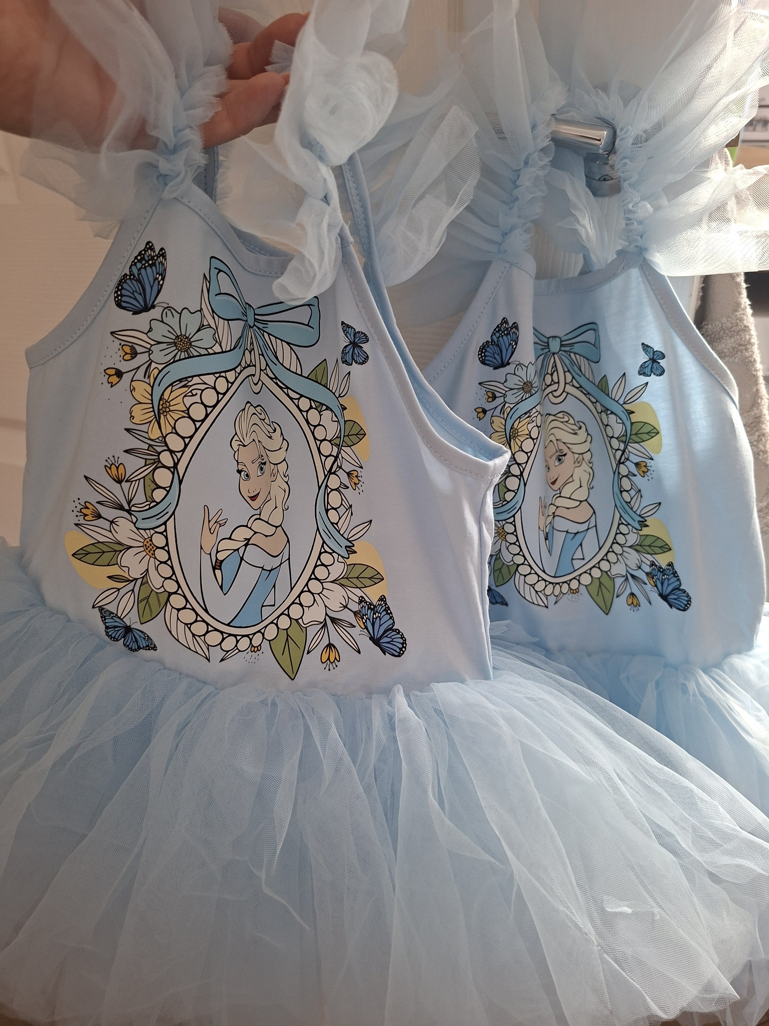 A white dress with a tulle skirt and a graphic print of a cartoon princess with blonde hair, blue eyes, and a light blue dress, surrounded by butterflies and flowers.
