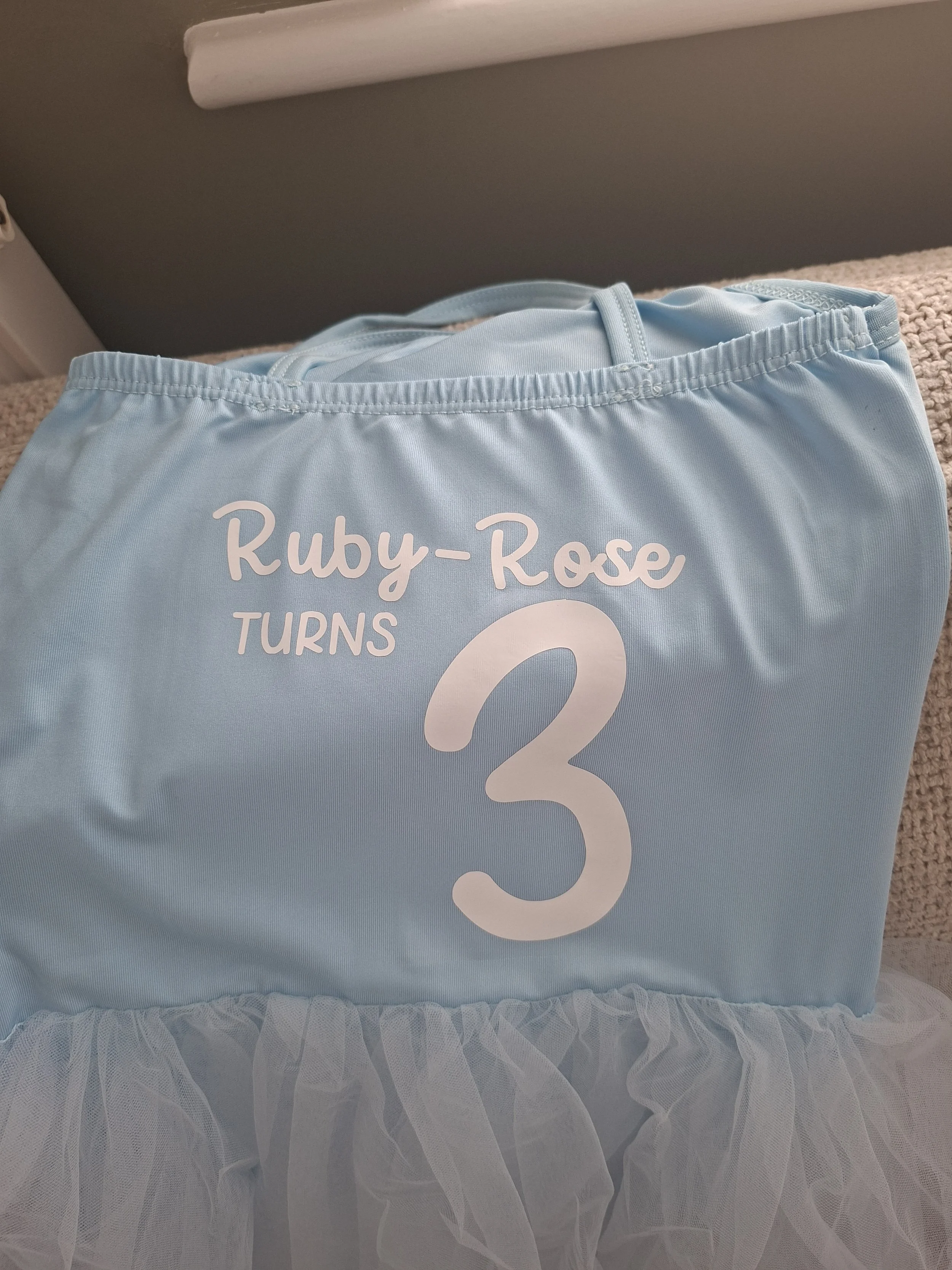 Light blue fabric with white text that says 'Ruby-Rose TURNS 2' and a large white number 2, with white ruffled tulle at the bottom.