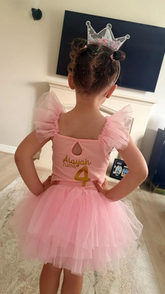 Back view of a young girl in a pink tutu dress with puffed sleeves, wearing a silver crown headband, and standing with hands on her hips in a living room.