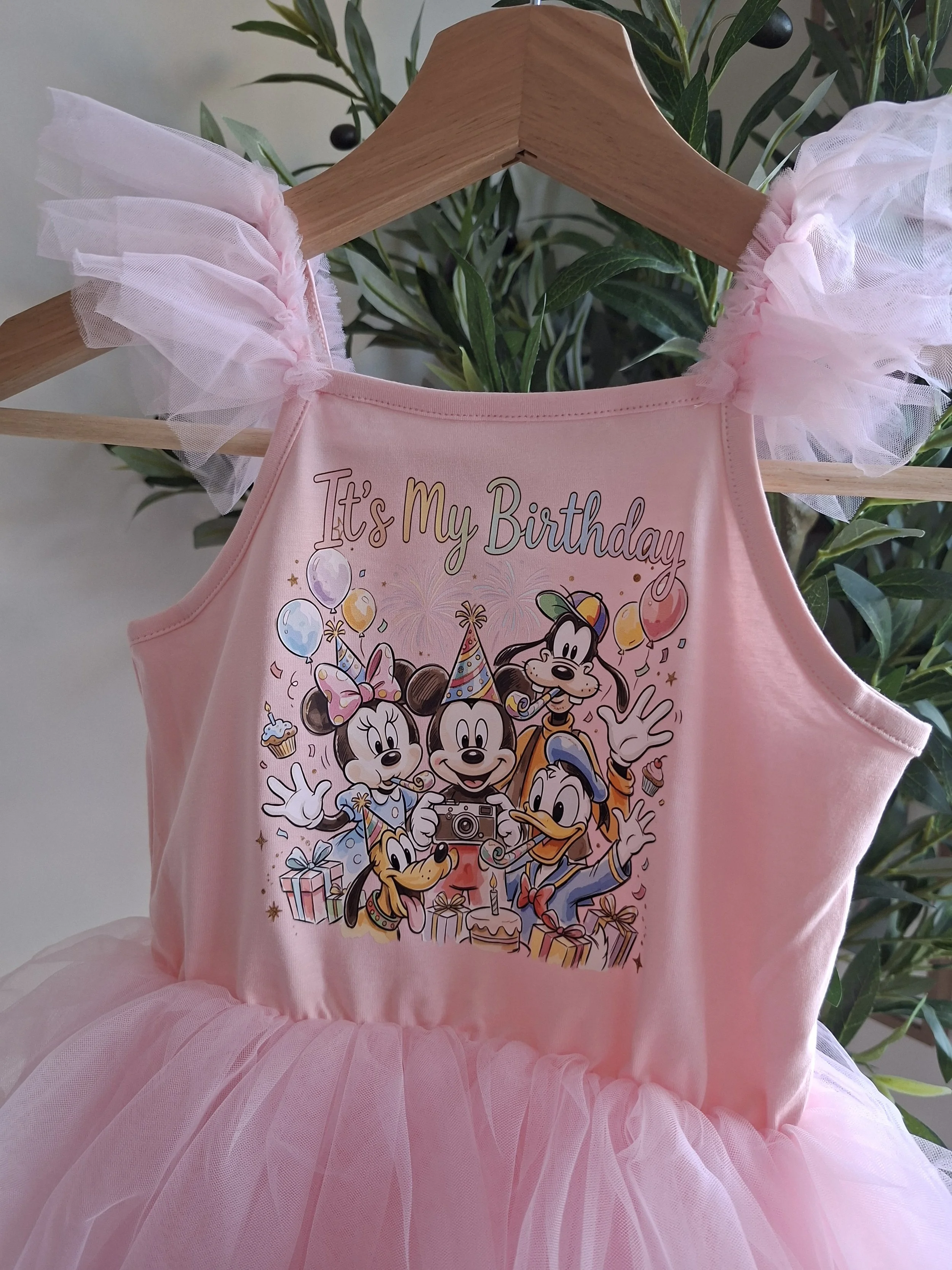 A pink birthday dress with a tulle tutu skirt and cartoon Disney characters Minnie Mouse, Mickey Mouse, Donald Duck, Daisy Duck, and Goofy, surrounded by balloons, confetti, and presents, hanging on a wooden hanger in front of a green leafy plant.